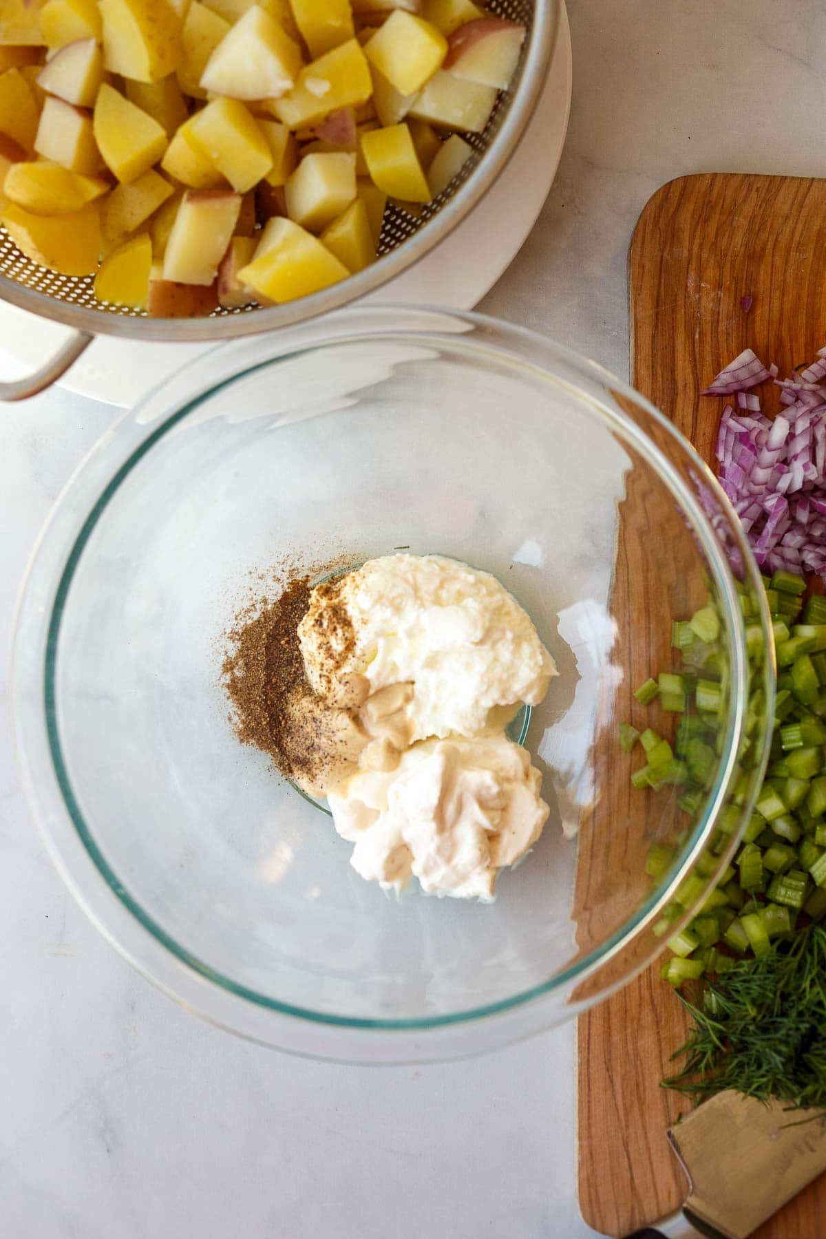 glass mixing bowl with potato salad dressing ingredients - greek yogurt, mayonnaise, pickle brine, dijon, salt, and pepper.