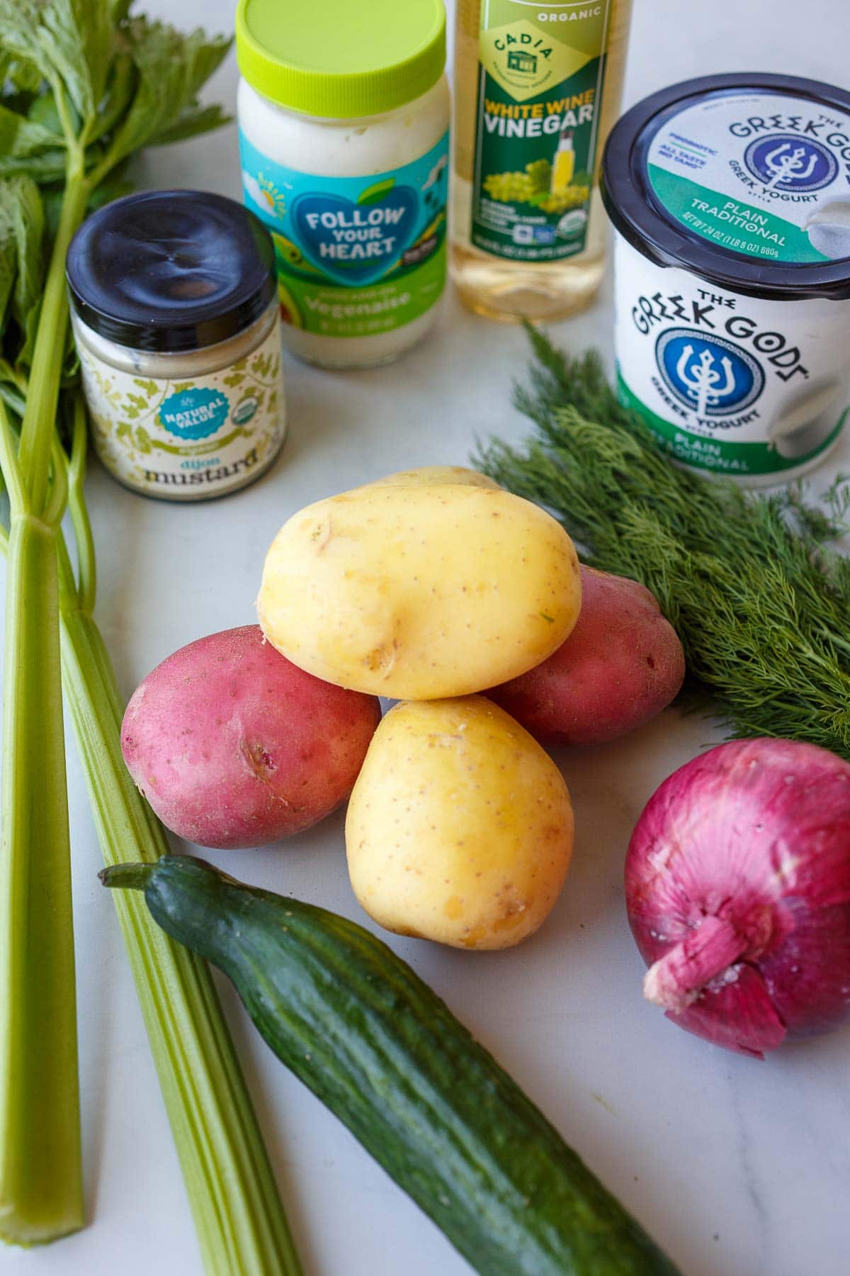 ingredients for potato salad laid out - potatoes, dill, onion, cucumber, celery, dijon, greek yogurt, vegenaise, white wine vinegar.