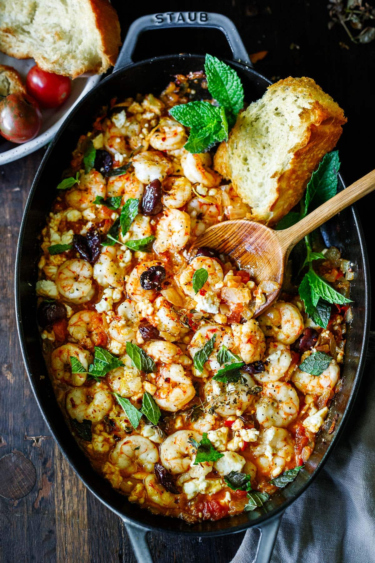 shrimp saganaki in ovenproof dish with wood serving spoon, garnished with mint and served with a crusty bread slice.