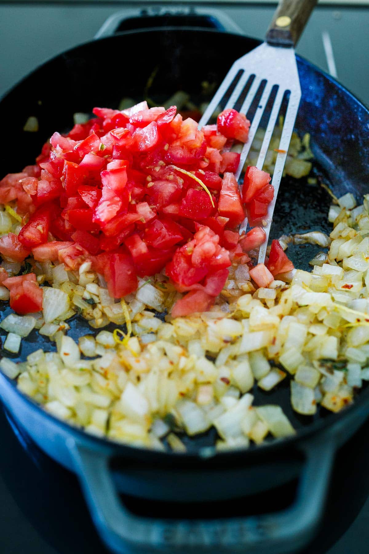 metal spatula stirring diced tomatoes into skillet with sauteed onions.