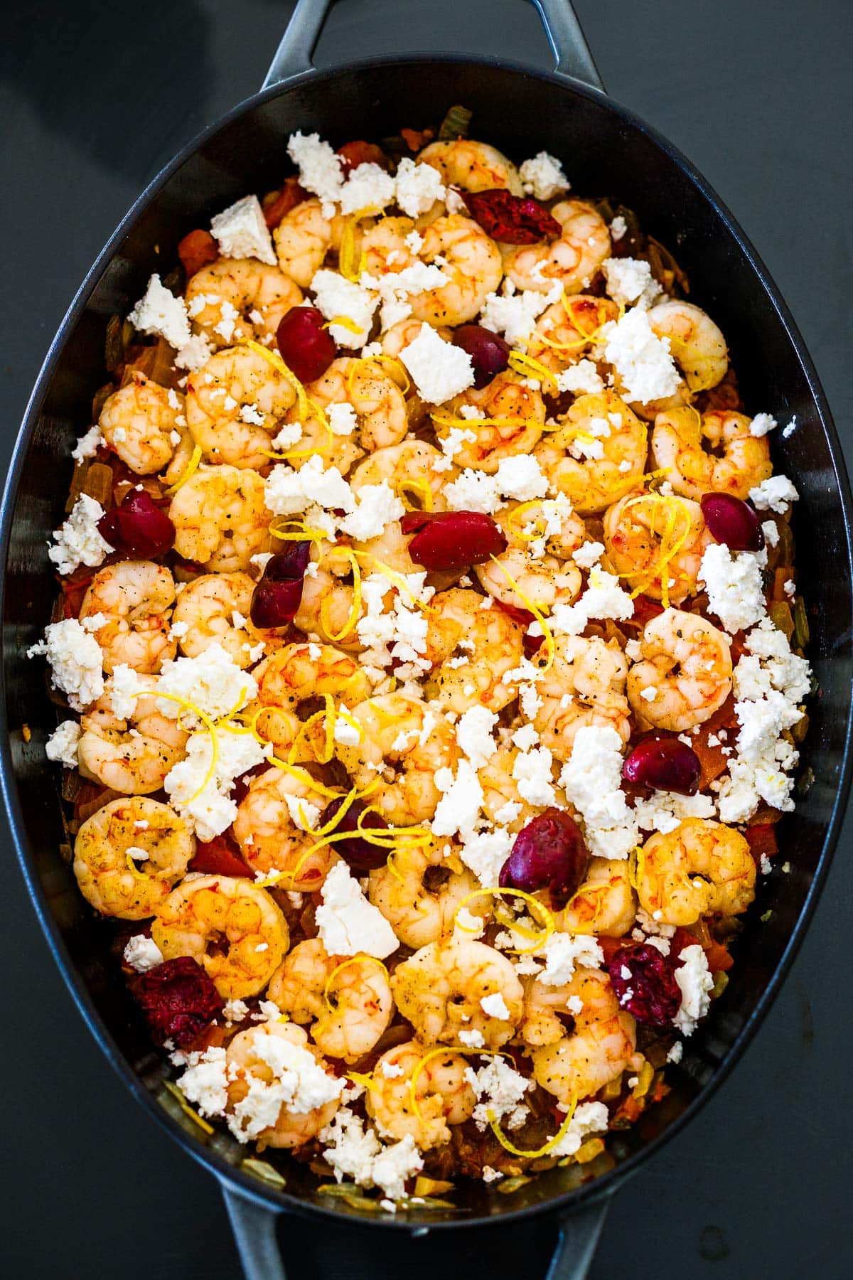 assembled, unbaked shrimp saganaki with crumbled feta, olives, lemon zest, and shrimp.