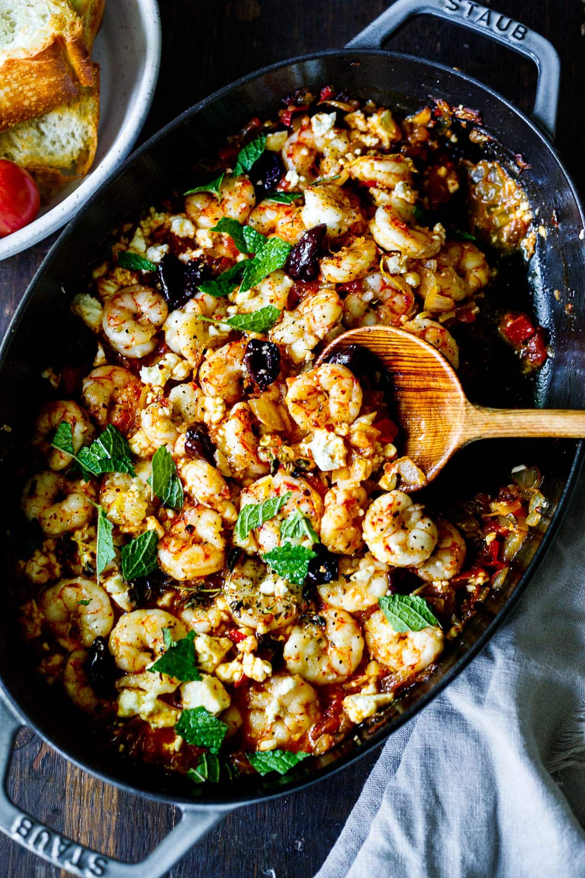 shrimp saganaki with olives in pan with wood spoon and fresh mint garnish.