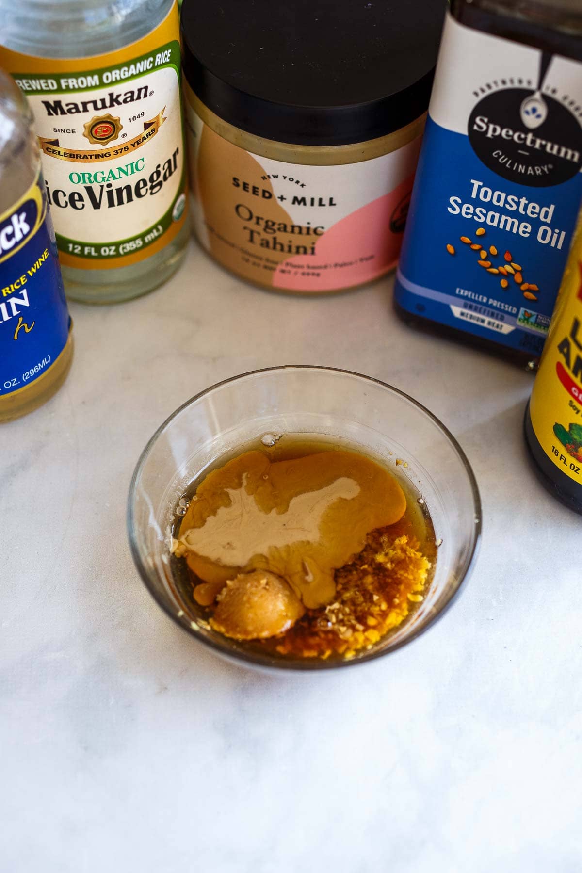 sauces, oil, and vinegar around small bowl with ingredients for sesame ginger dressing. 