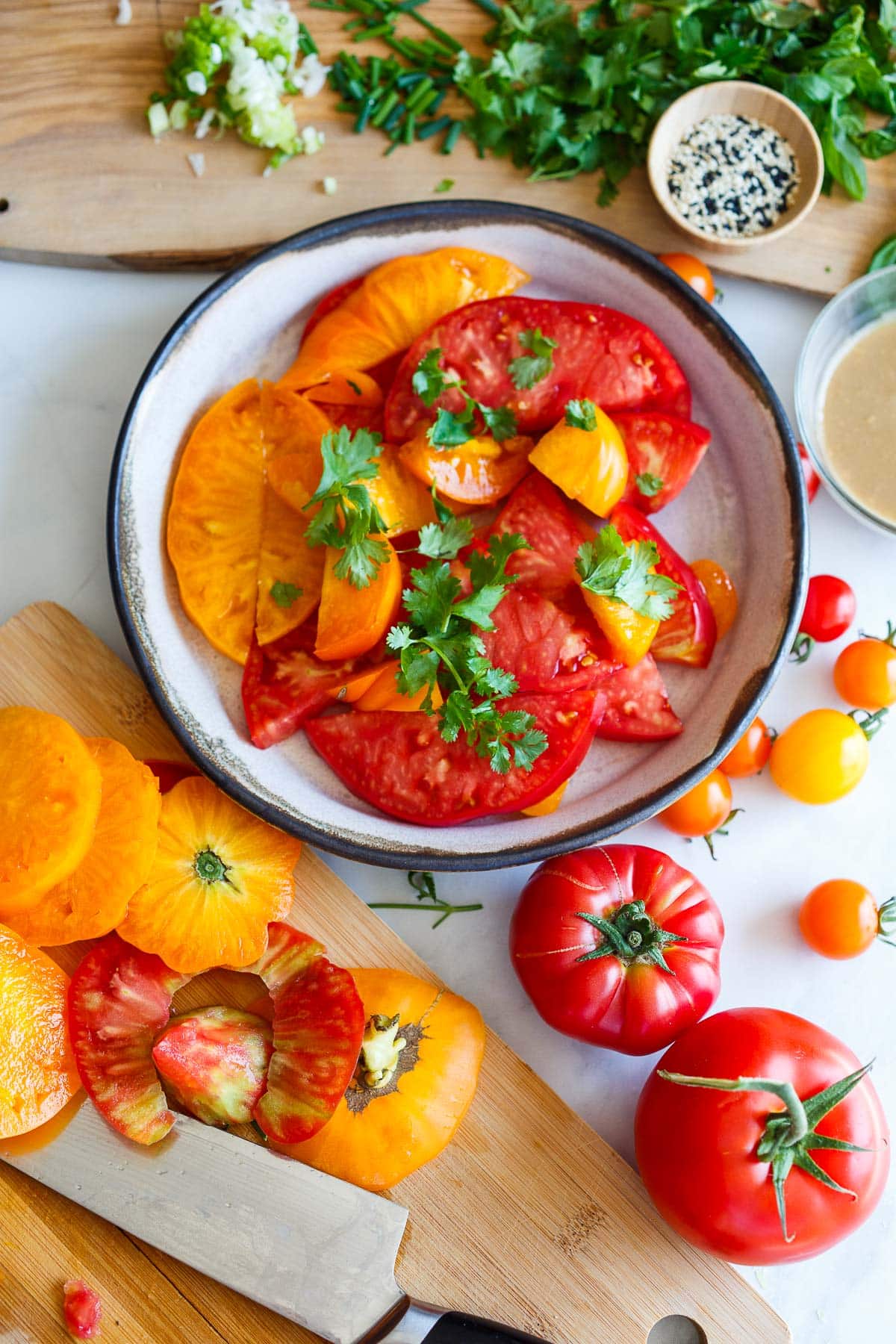 serving bowl with sliced heirloom tomatoes topped with fresh herbs next to wood boards with chopped herbs, and sliced heirloom tomatoes. 