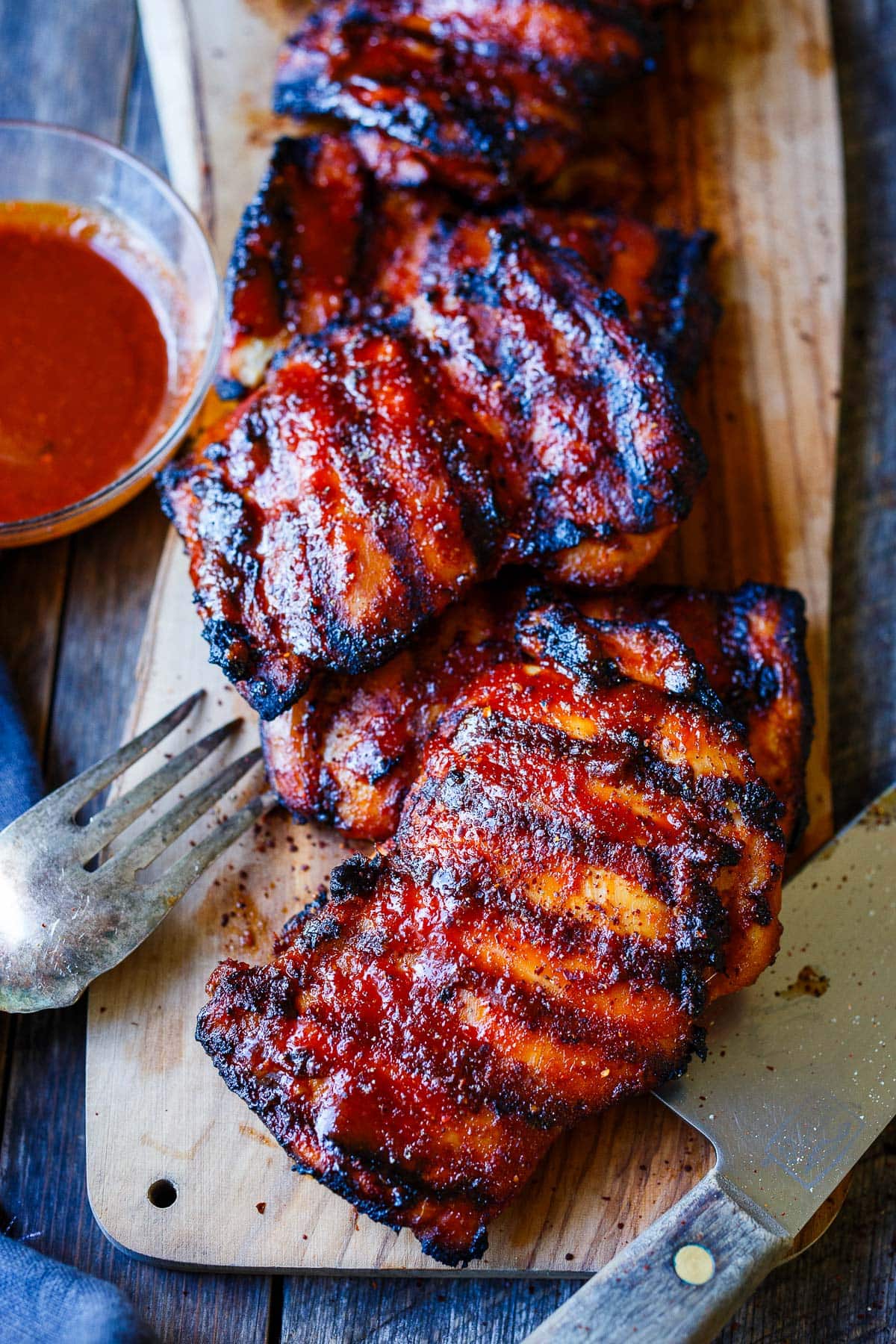 wood plank with grilled Korean bbq chicken laid in a row next to bowl of bbq sauce. 