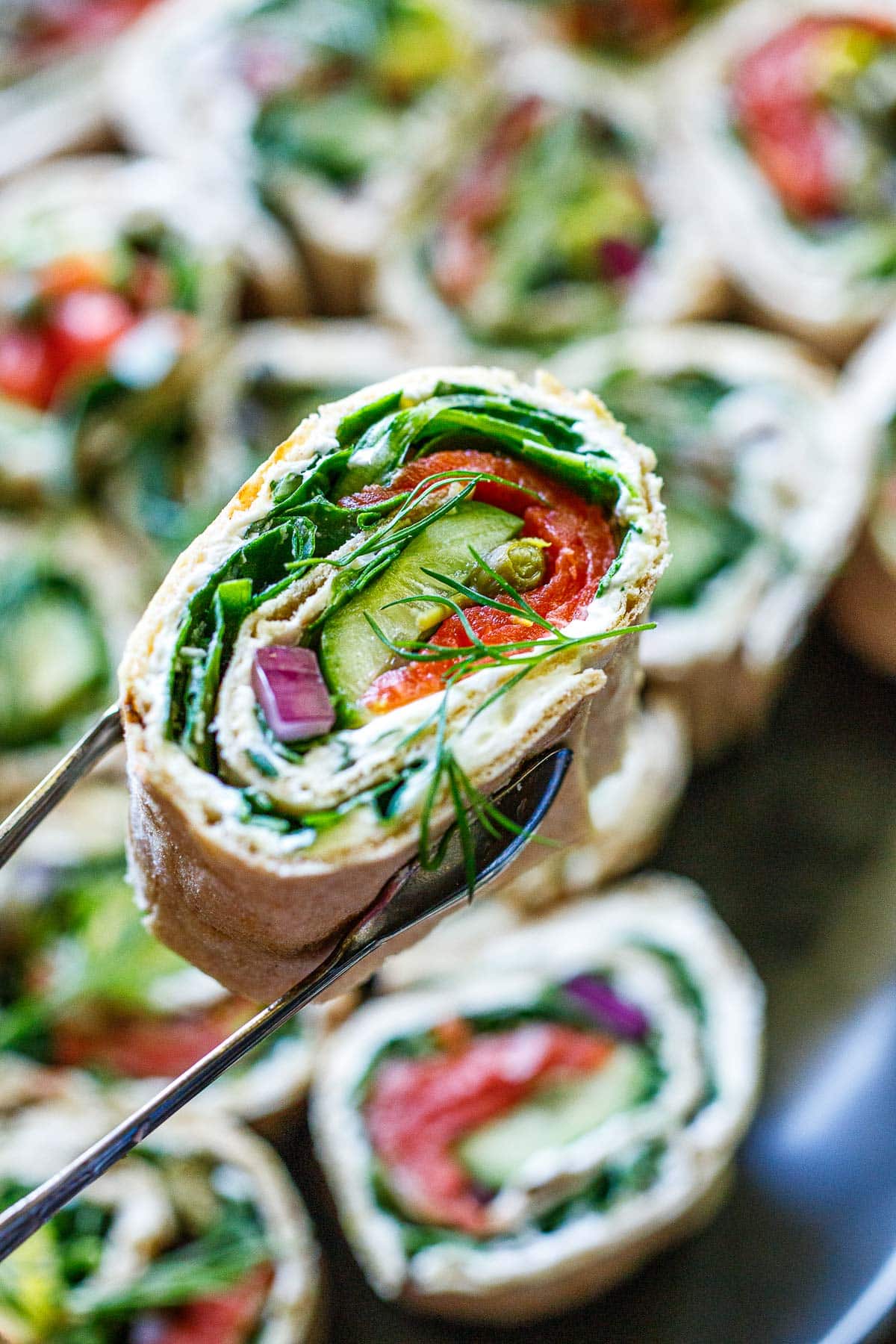 tongs holding lavash wrap filled with cream cheese, smoked salmon, avocado, cucumber, red onion, capers, and dill. 