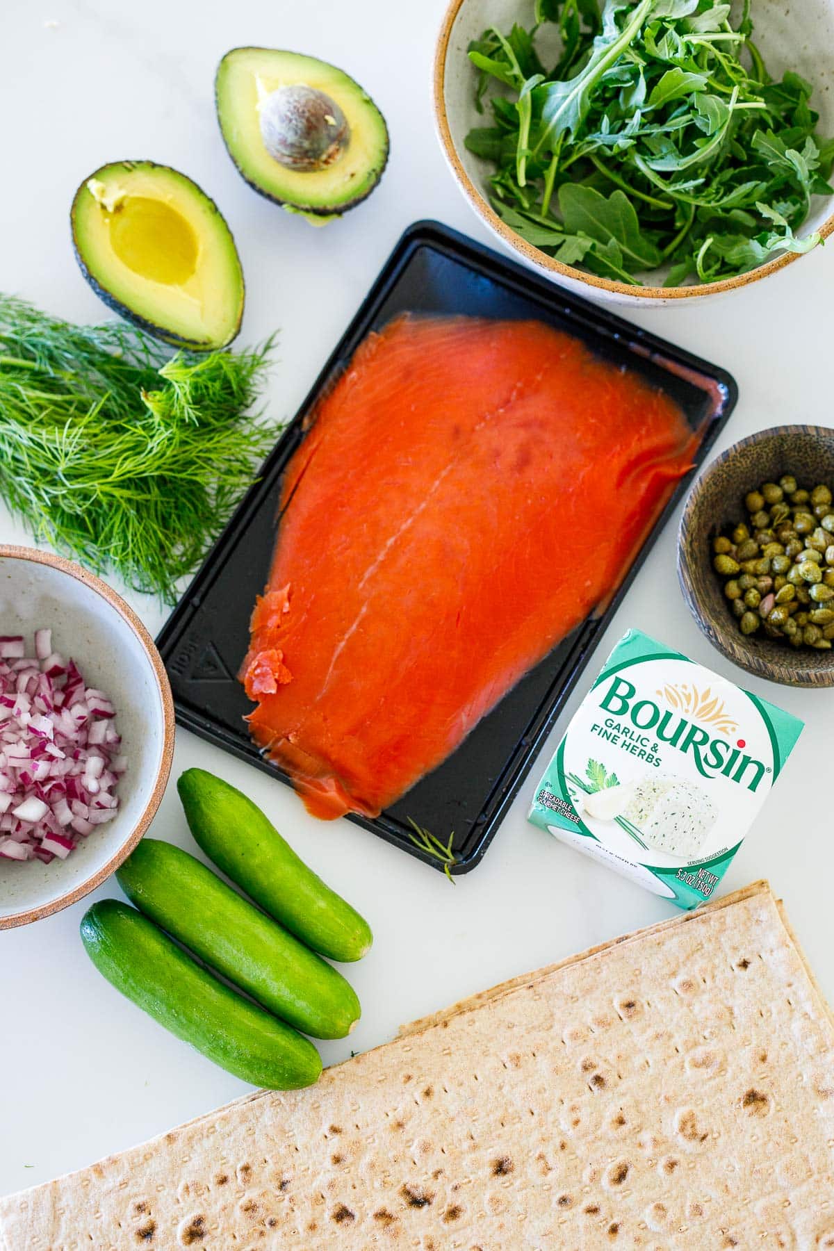 ingredients laid out for lavash wraps- bowl of arugula, halved avocado, fresh dill bunch, smoked salmon, bowl of capers, bowl of diced red onion, boursin garlic herb cream cheese, turkish cucumbers, and lavash bread. 