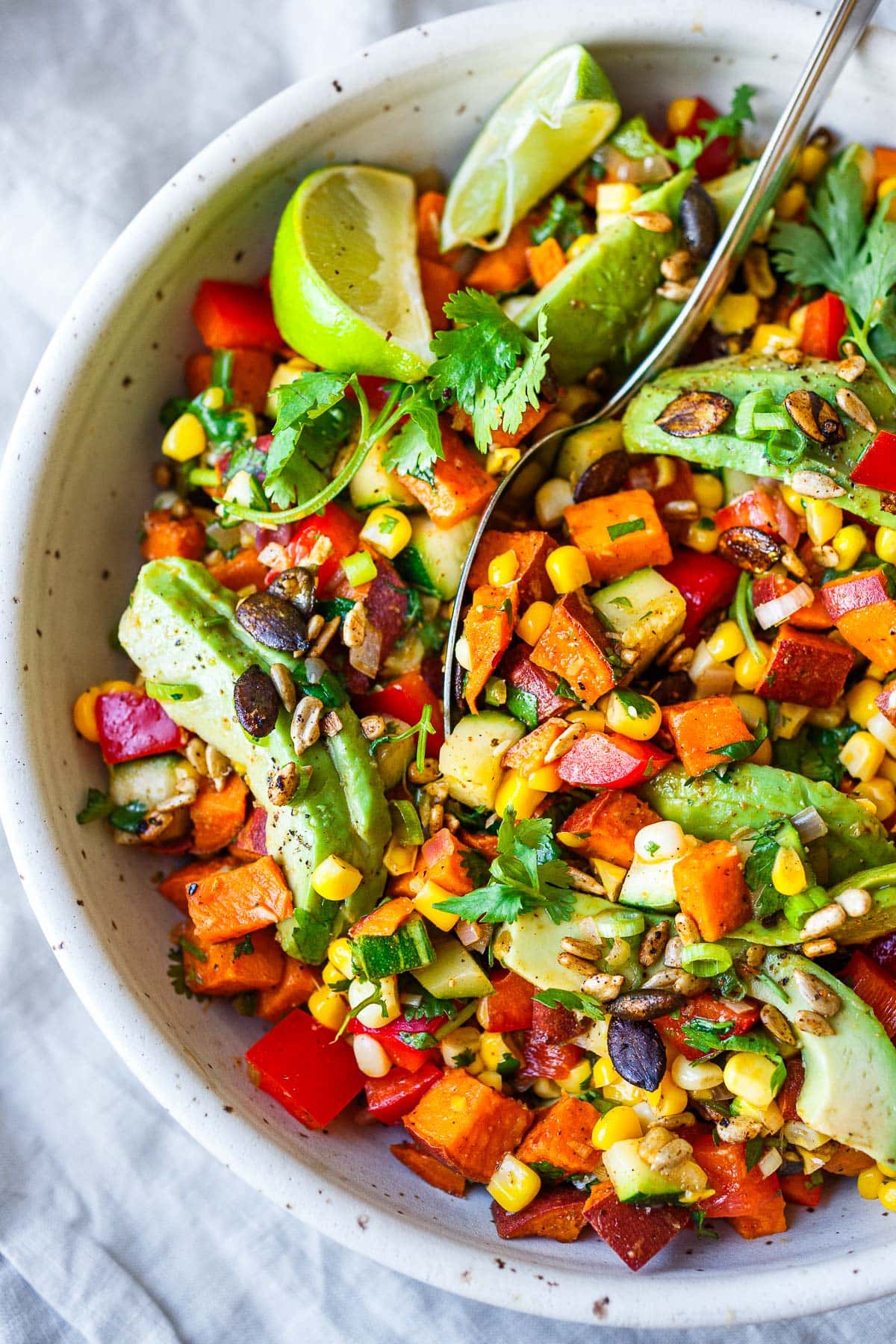 salad bowl with sweet potato salad and spoon garnished with sliced avocado, cilantro, and lime wedges.