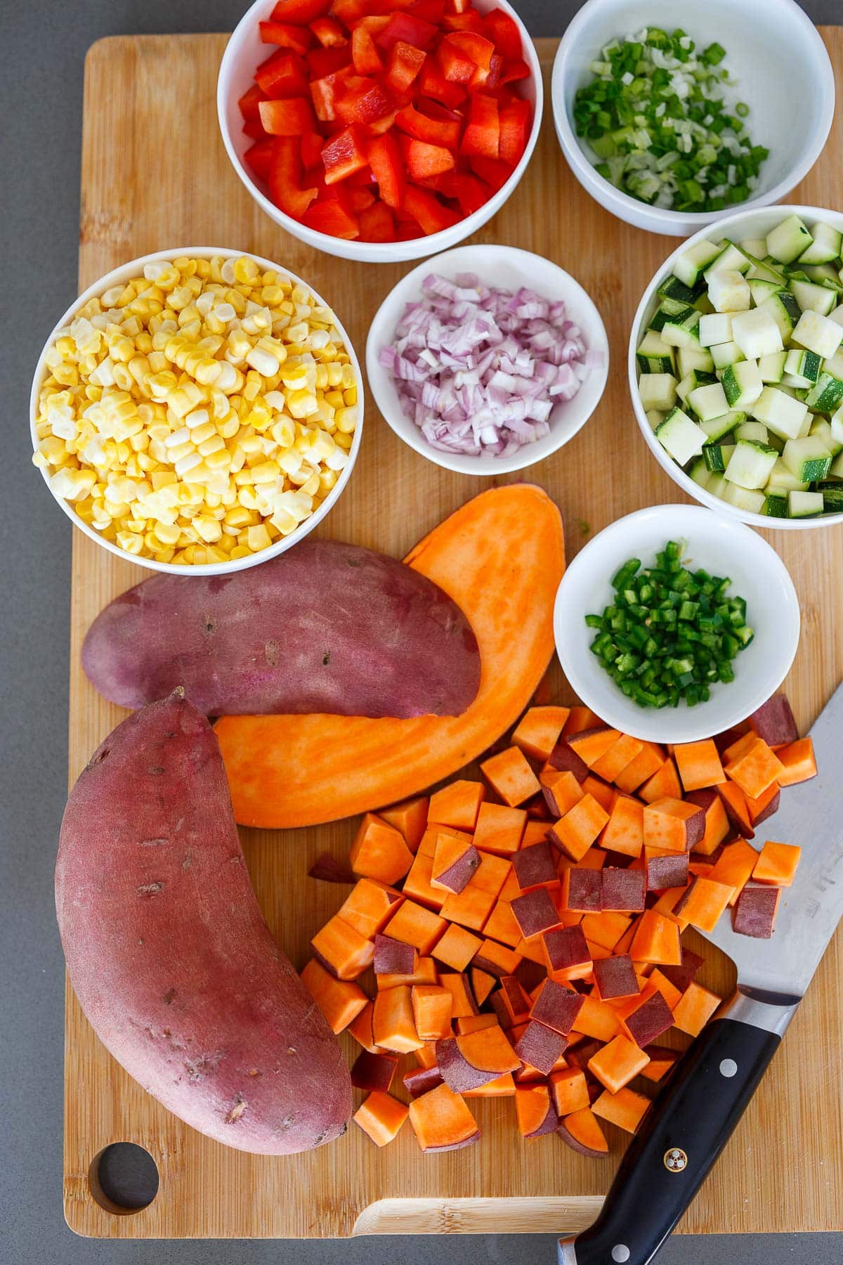 wood cutting board and sharp knife with diced sweet potatoes and small bowls with corn, bell pepper, green onion, shallot, zucchini, and jalapeño.