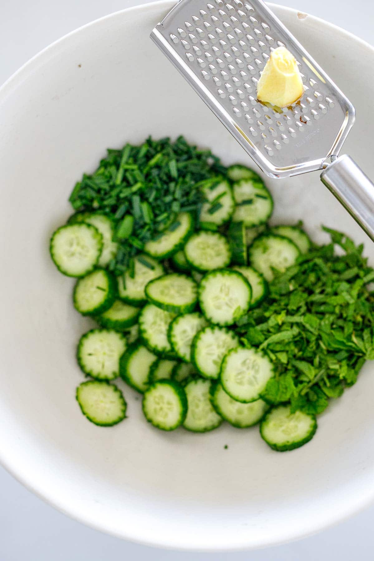 large salad bowl with sliced cucumbers and herbs and ginger on microplane grater above bowl. 