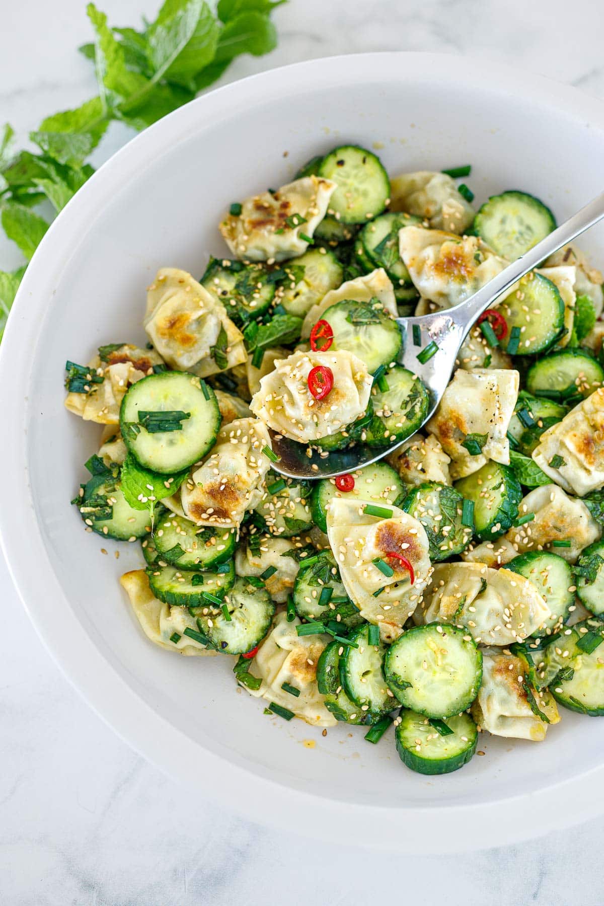 wonton salad in bowl with spoon with sliced cucumbers, red chilis, and toasted sesame seeds. 