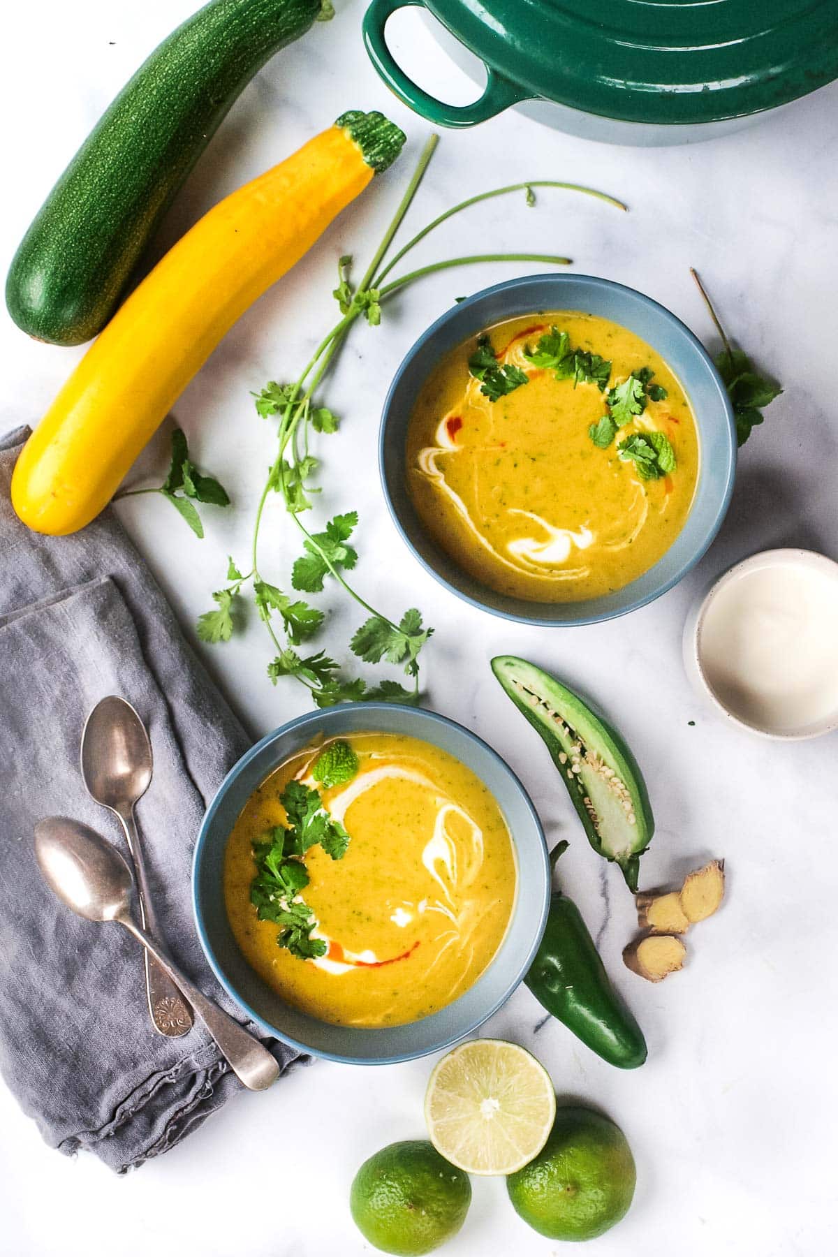 two bowls of curried zucchini soup garnished with coconut milk and fresh herbs, next to jalapeño, ginger, herbs, spoons, napkins, and limes. 