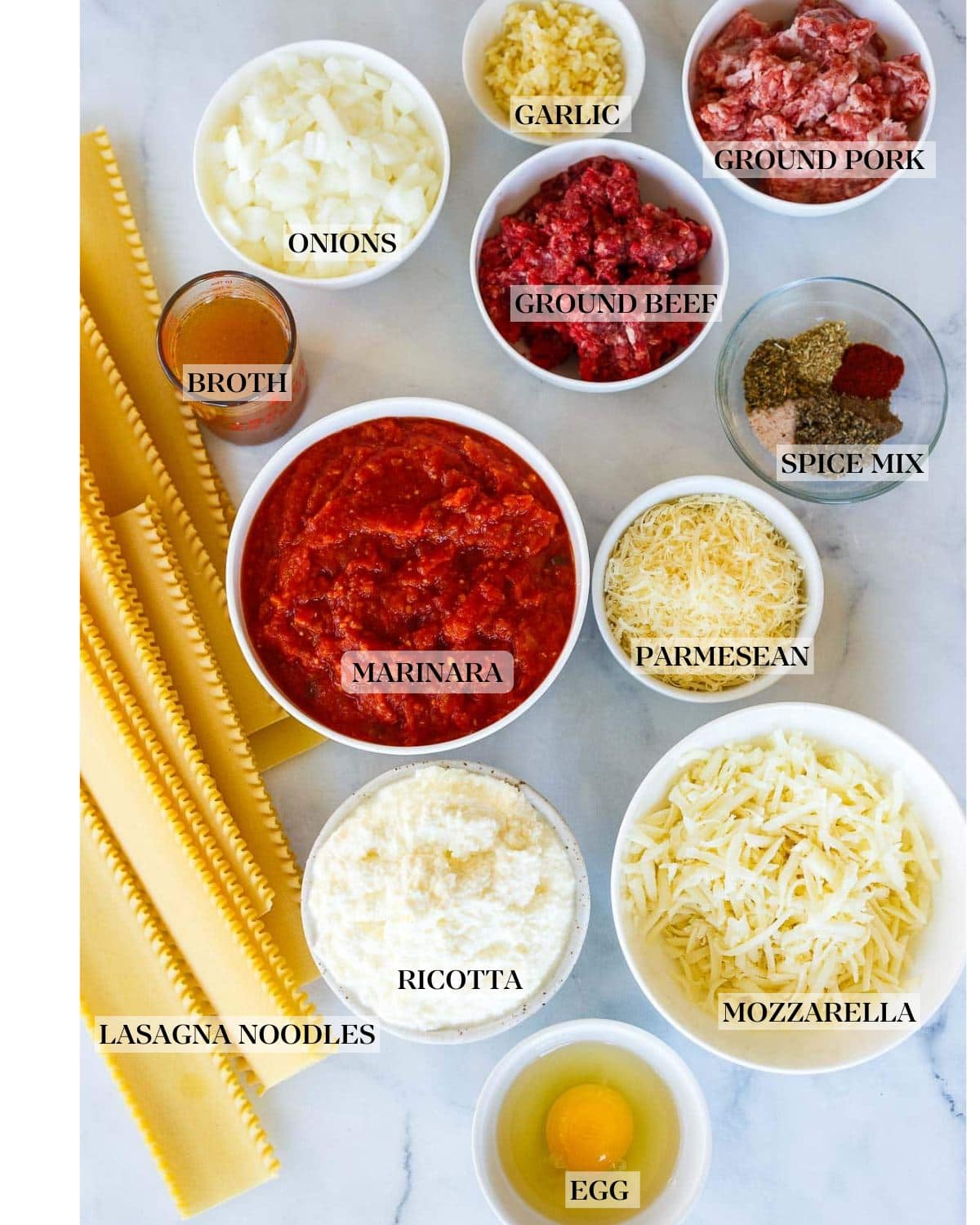 ingredients for traditional lasagna laid out and labeled - garlic, onions, broth, ground pork, ground beef, spice mix, parmesan, marinara, mozzarella, ricotta, lasagna noodles, egg.