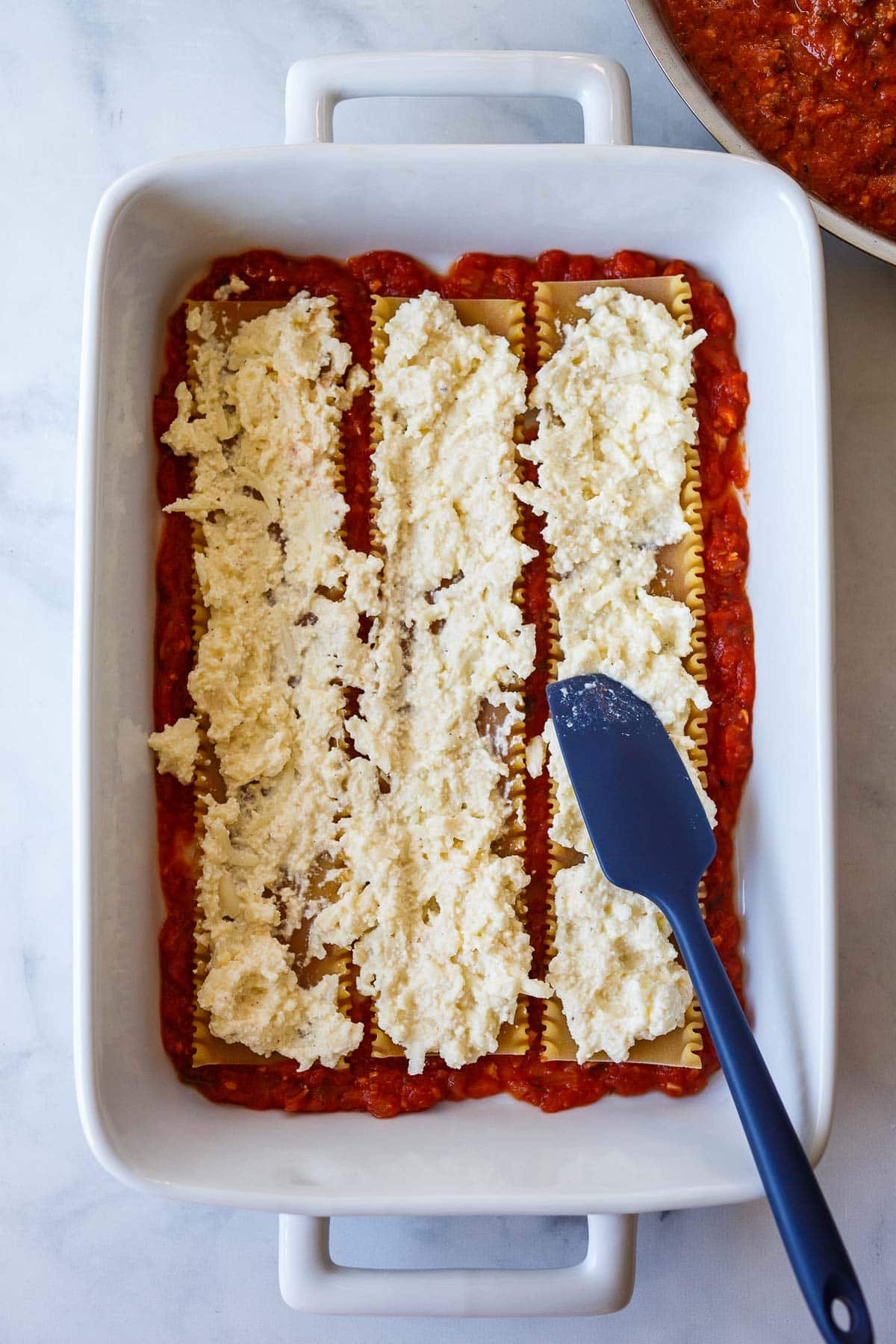 baking dish with spatula spreading ricotta cheese mixture over lasagna noodles.