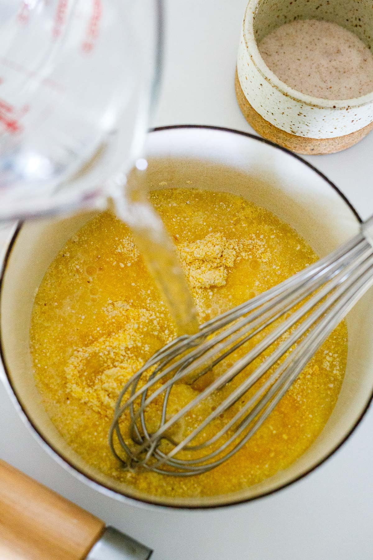 measuring cup pouring water into pot with cornmeal and whisk.