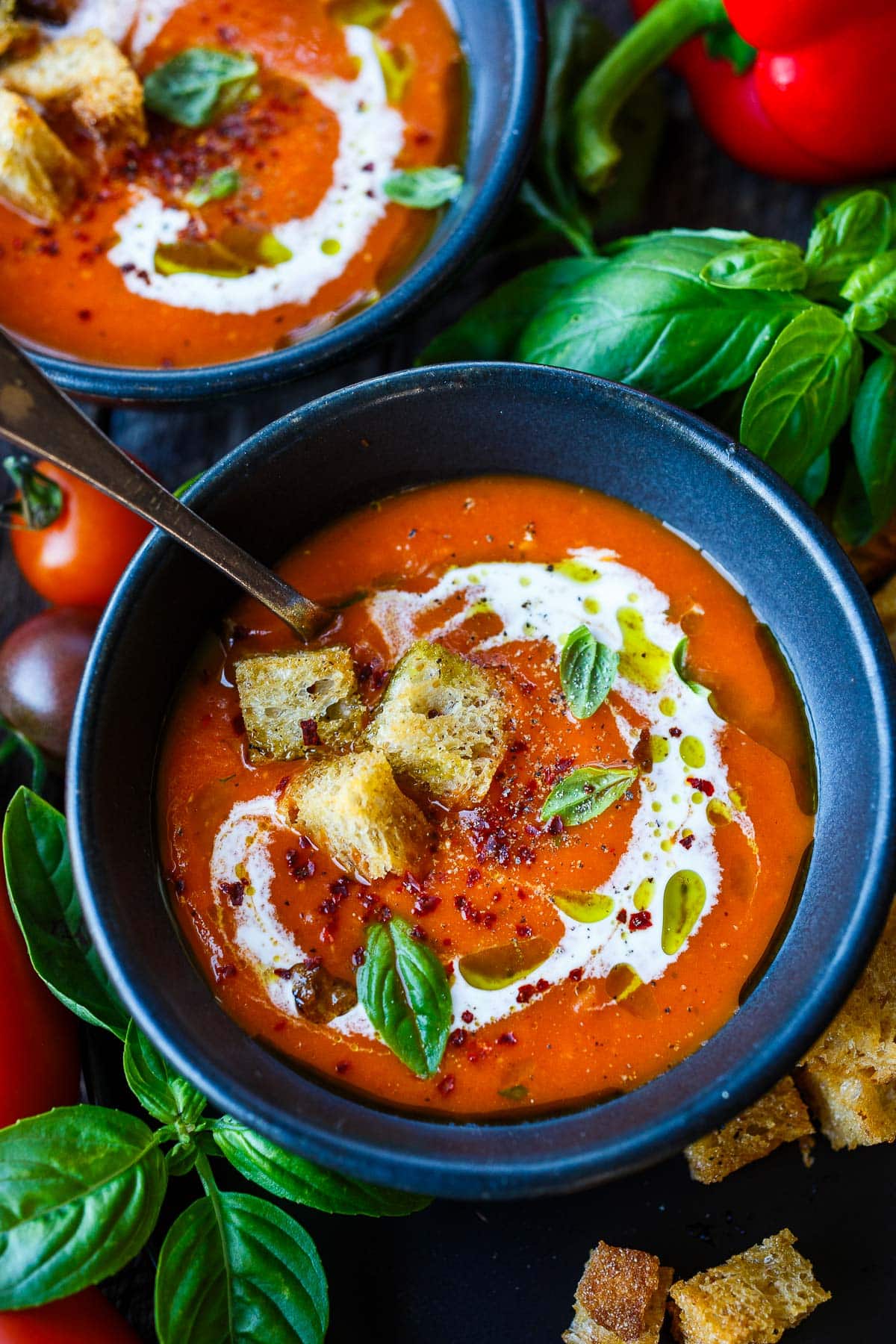 bowl of roasted red pepper tomato soup with spoon and swirl of yogurt, topped with basil oil, fresh basil leaves, and croutons.