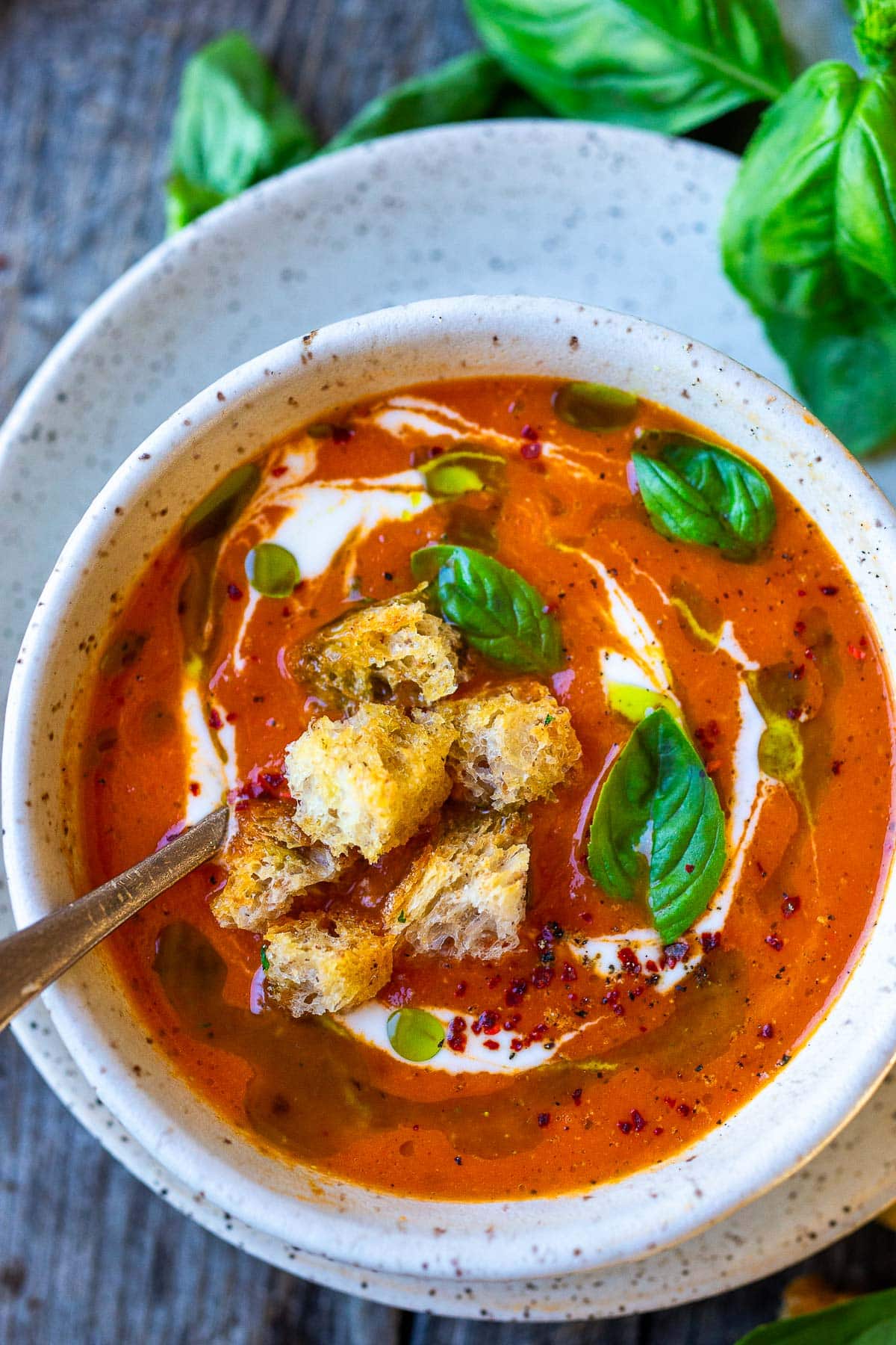soup bowl and spoon with roasted red pepper and tomato soup garnished with swirl of coconut milk, fresh basil leaves, aleppo, oil, and croutons. 