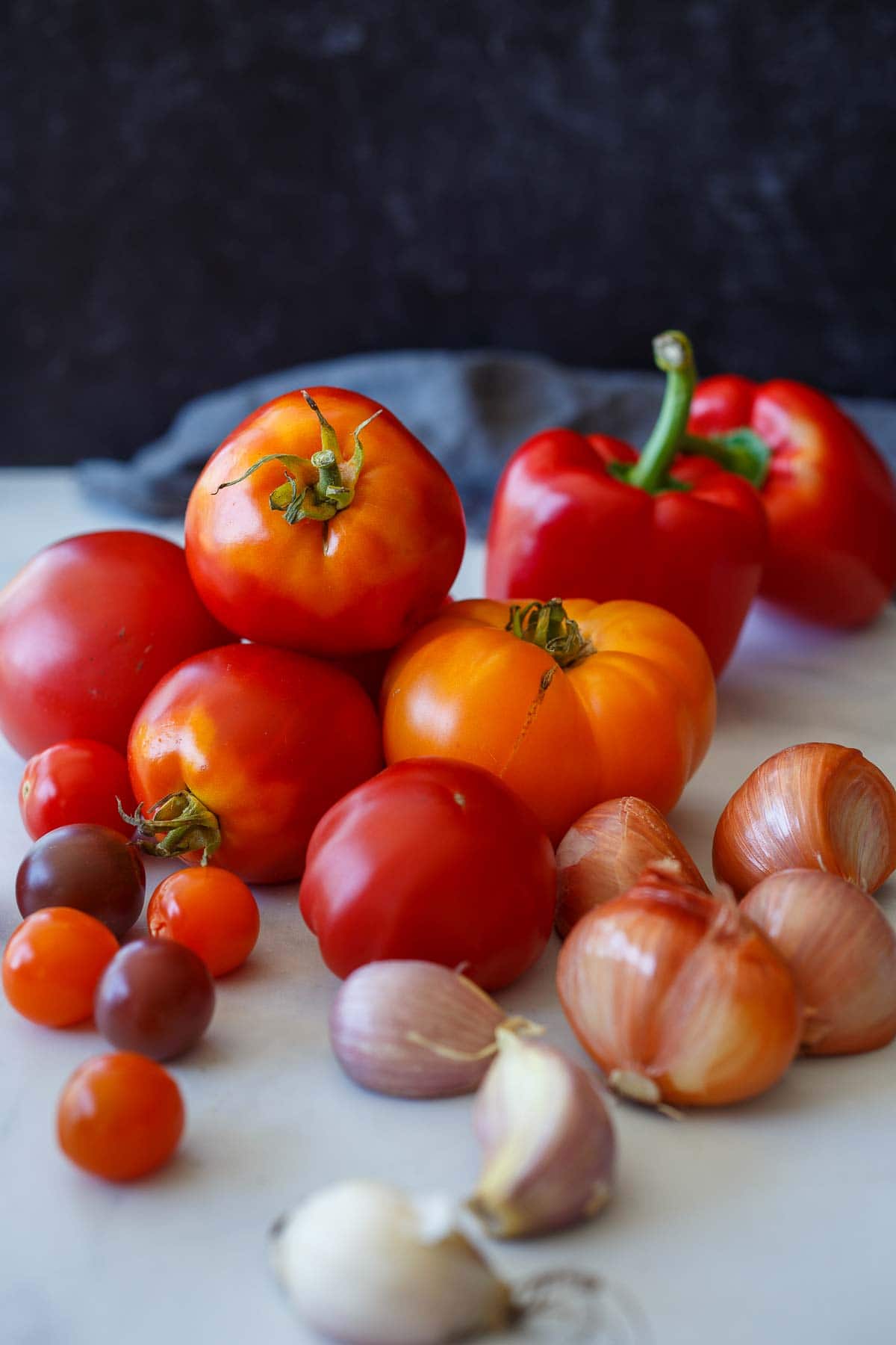 fresh ingredients for roasted red pepper tomato soup laid out - red bell peppers, medley of heirloom tomatoes and cherry tomatoes, shallots, and garlic. 