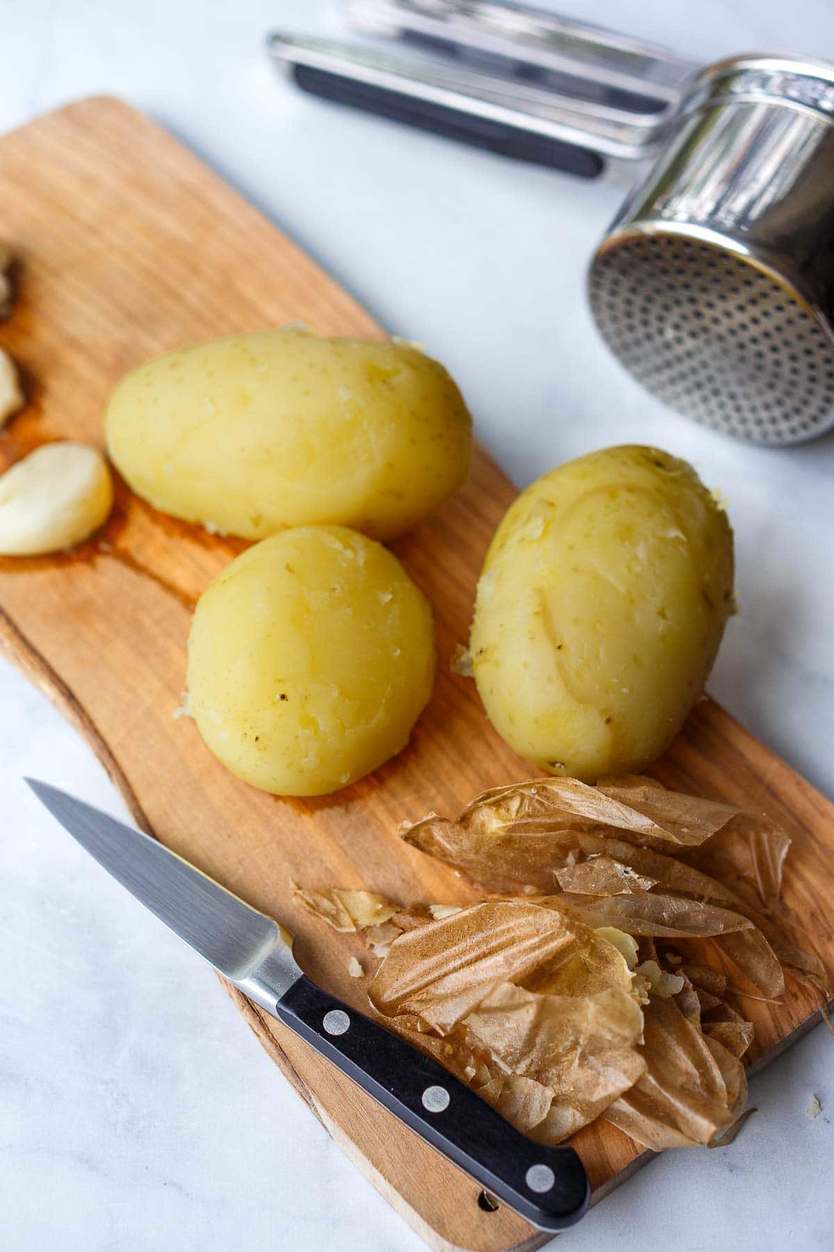 wood board with coiled potatoes peeled with sharp knife, skins to the side, with potato ricer next to the board. 