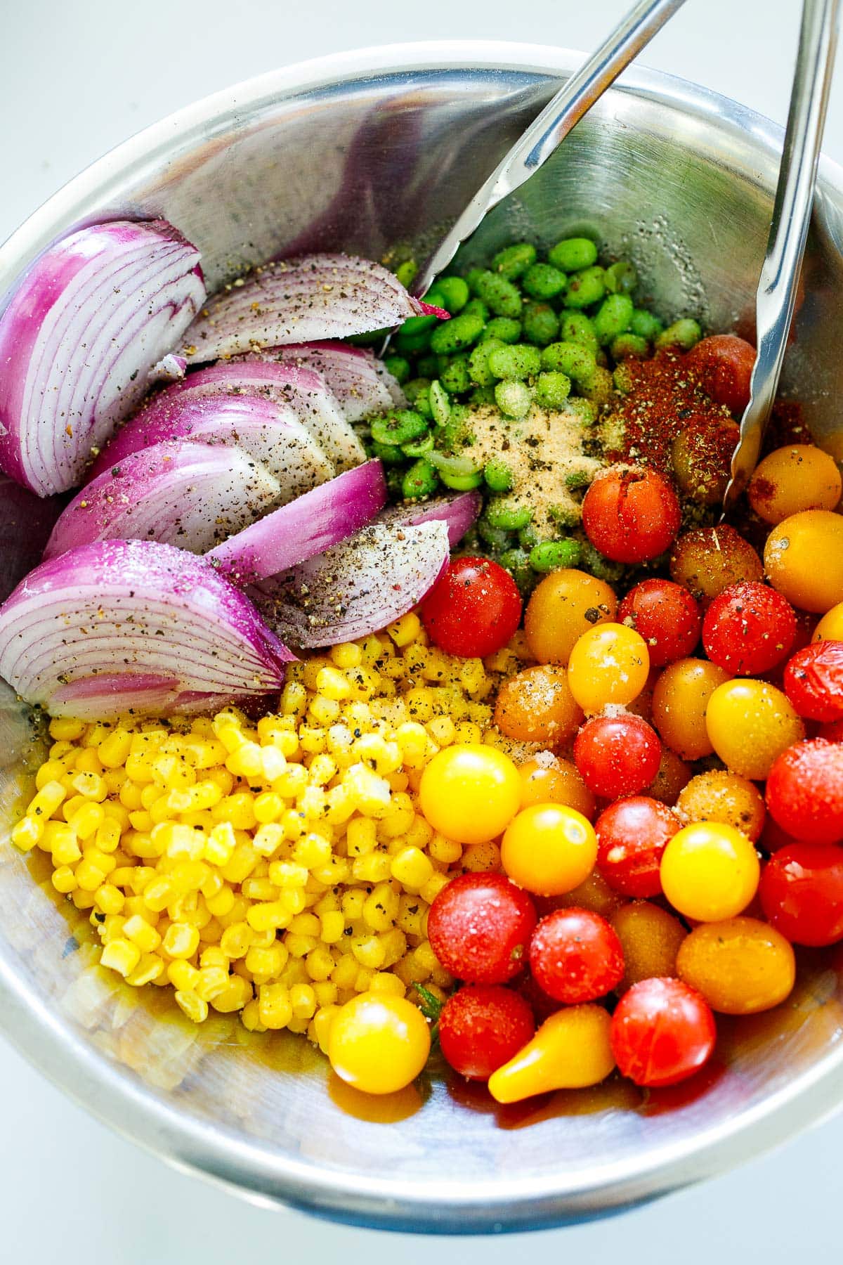 mixing bowl with succotash components - corn, cherry tomatoes, red onion, edamame, spices, tongs. 