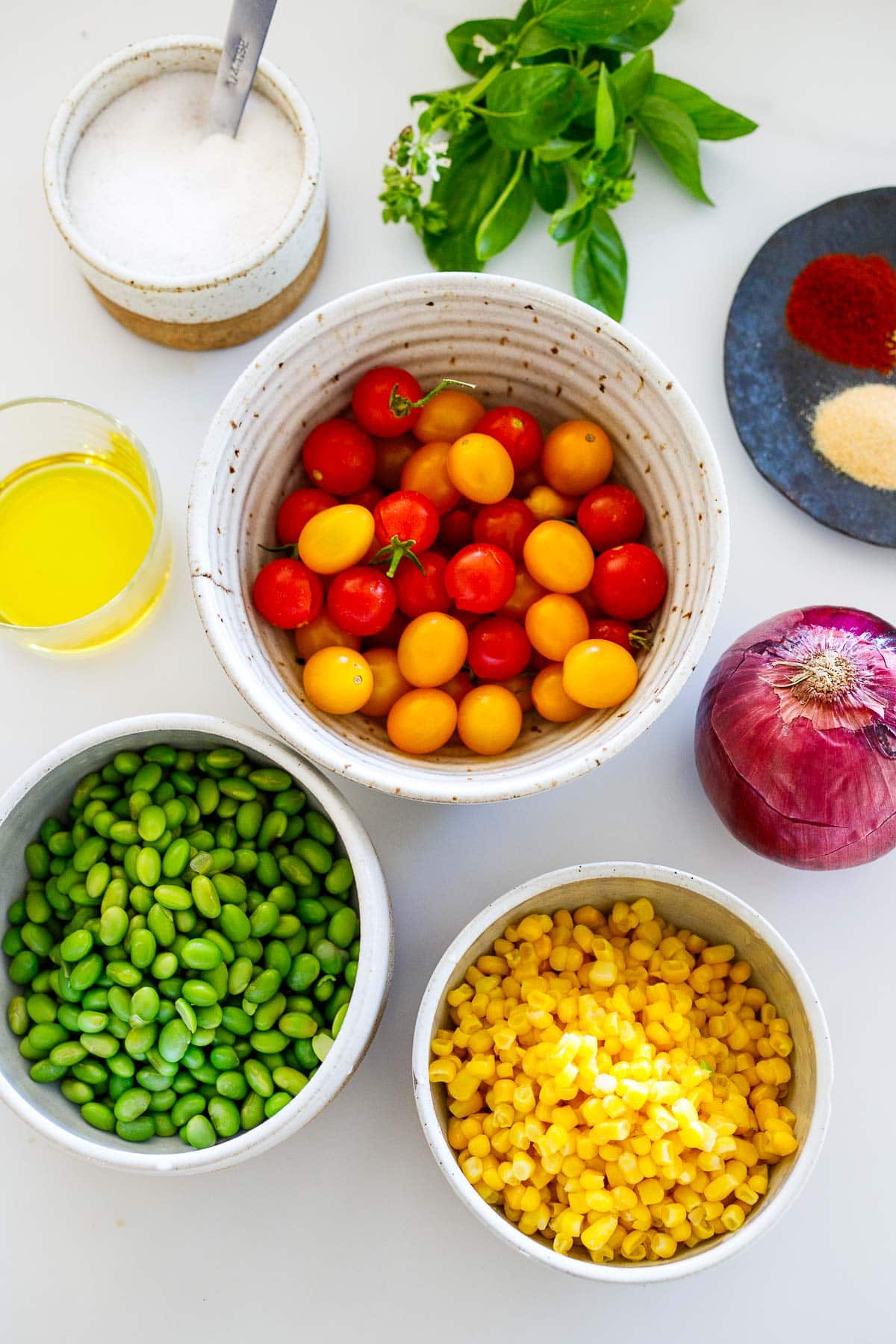 ingredients for sheet-pan succotash laid out - dish with spices, salt, glass of olive oil, bowl of cherry tomatoes, red onion, edamame, corn. 