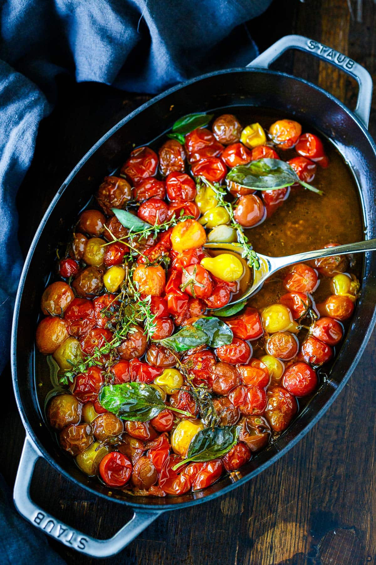 spoon lifting roasted tomatoes from tomato confit in oval dish with thyme and basil.