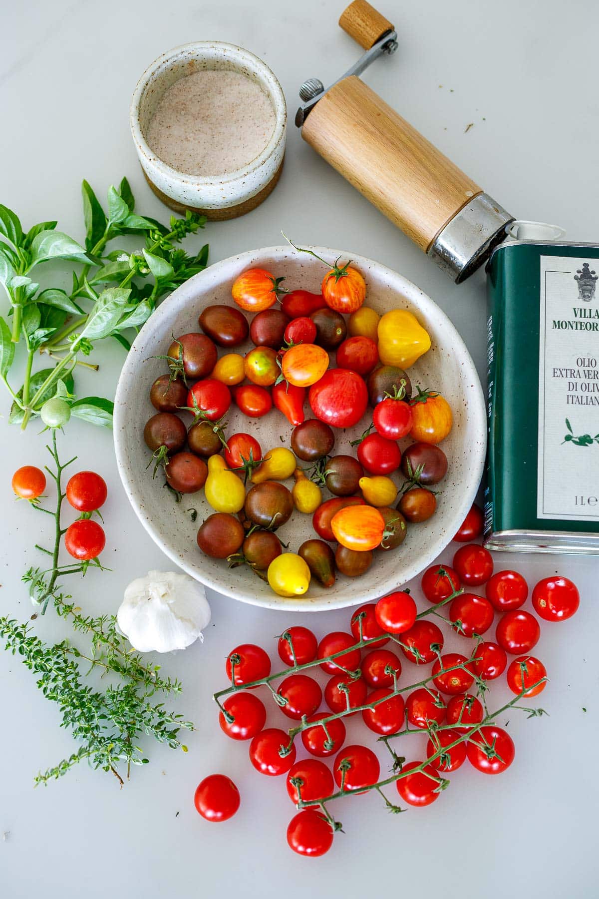 ingredients laid out for tomato confit - salt, pepper shaker, olive oil, bowl of mixed cherry tomatoes, basil, thyme, garlic. 