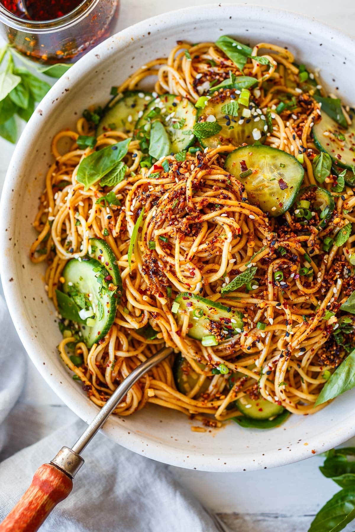 cold noodle salad in large serving bowl with utensil, topped with sliced cucumbers, mint, basil, green onions, sesame seeds, and chili crisp.