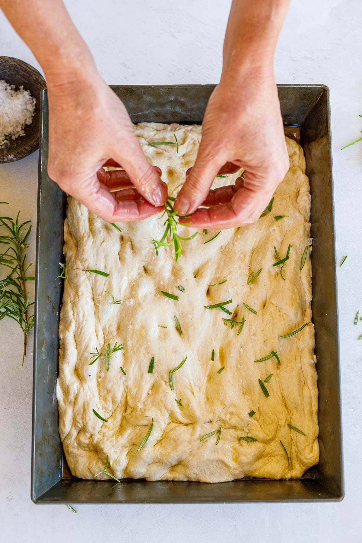 hands breaking apart fresh rosemary to scatter over focaccia.
