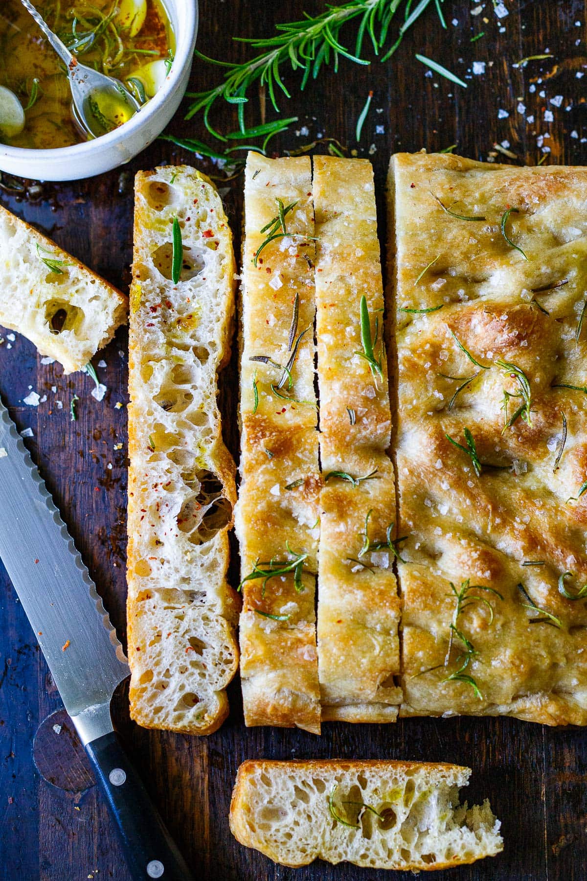 baked focaccia bread with rosemary and salt on wood board with sharp and thinly sliced pieces.