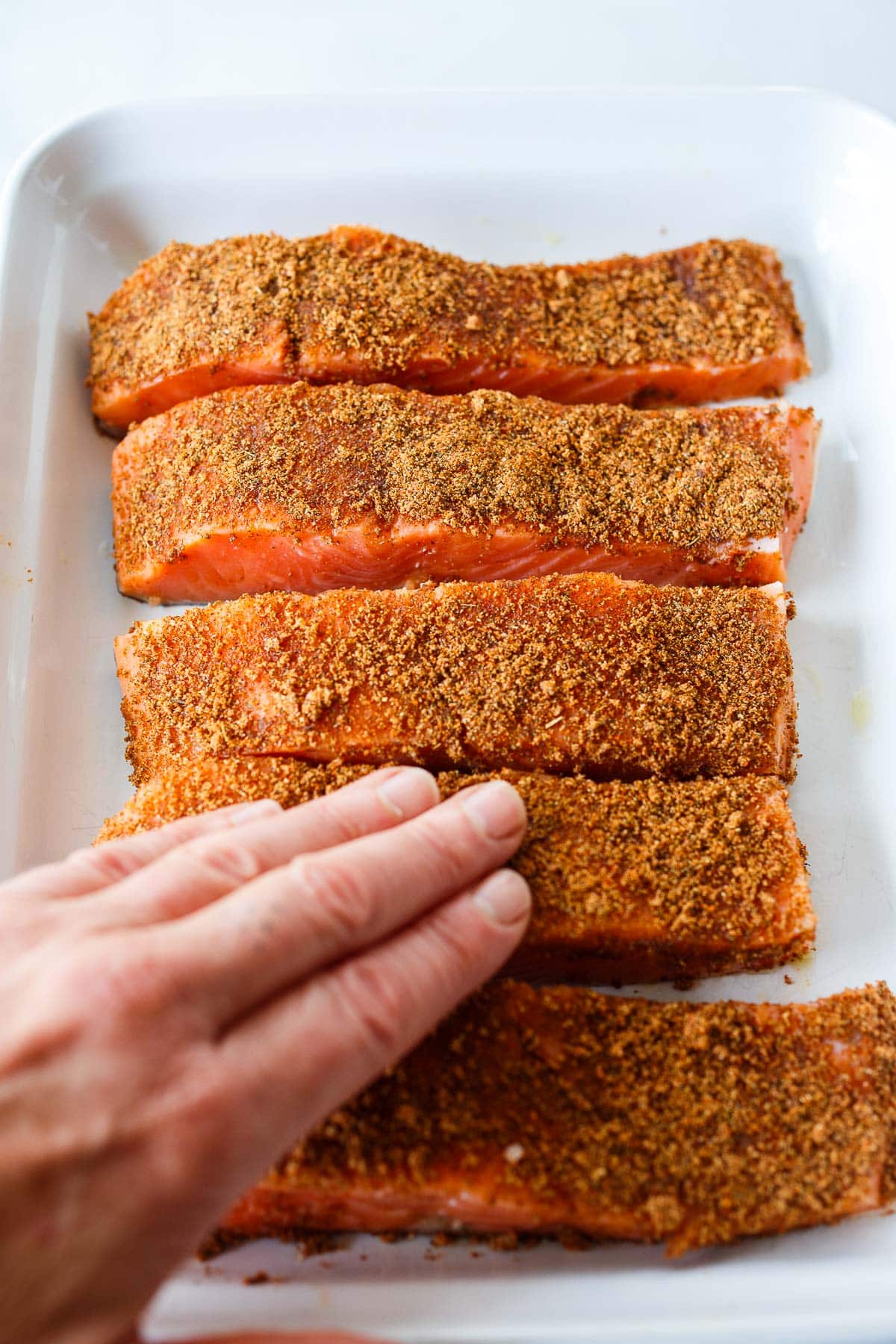 salmon filets in a tray with cajun seasoning spread over top and a hand pressing spices into the filets.