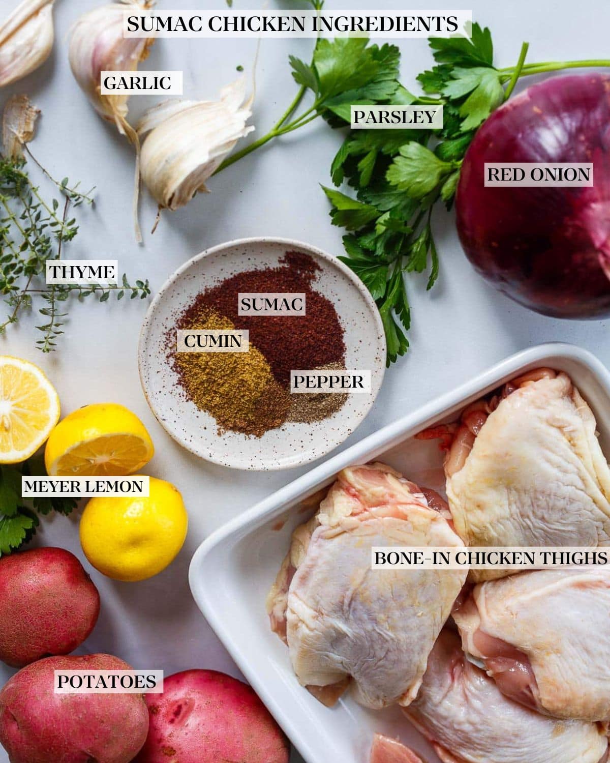 ingredients for roasted chicken with sumac - red onion, fresh parsley and thyme, garlic cloves, bowl of spices, lemon halves, red potatoes, and tray of chicken thighs.