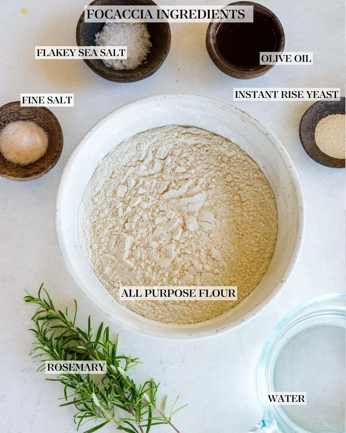 large mixing bowl with flour, next to small bowls with salt, oil, and yeast, fresh rosemary, and measuring cup with water.