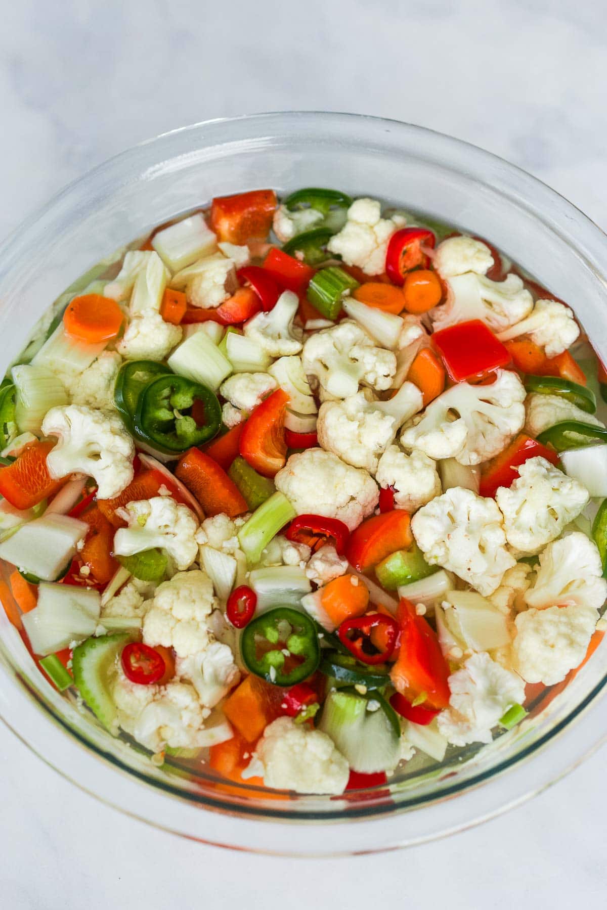 glass mixing bowl with bite size chopped veggies like cauliflower, bell peppers, chilis, and carrots, covered with water. 