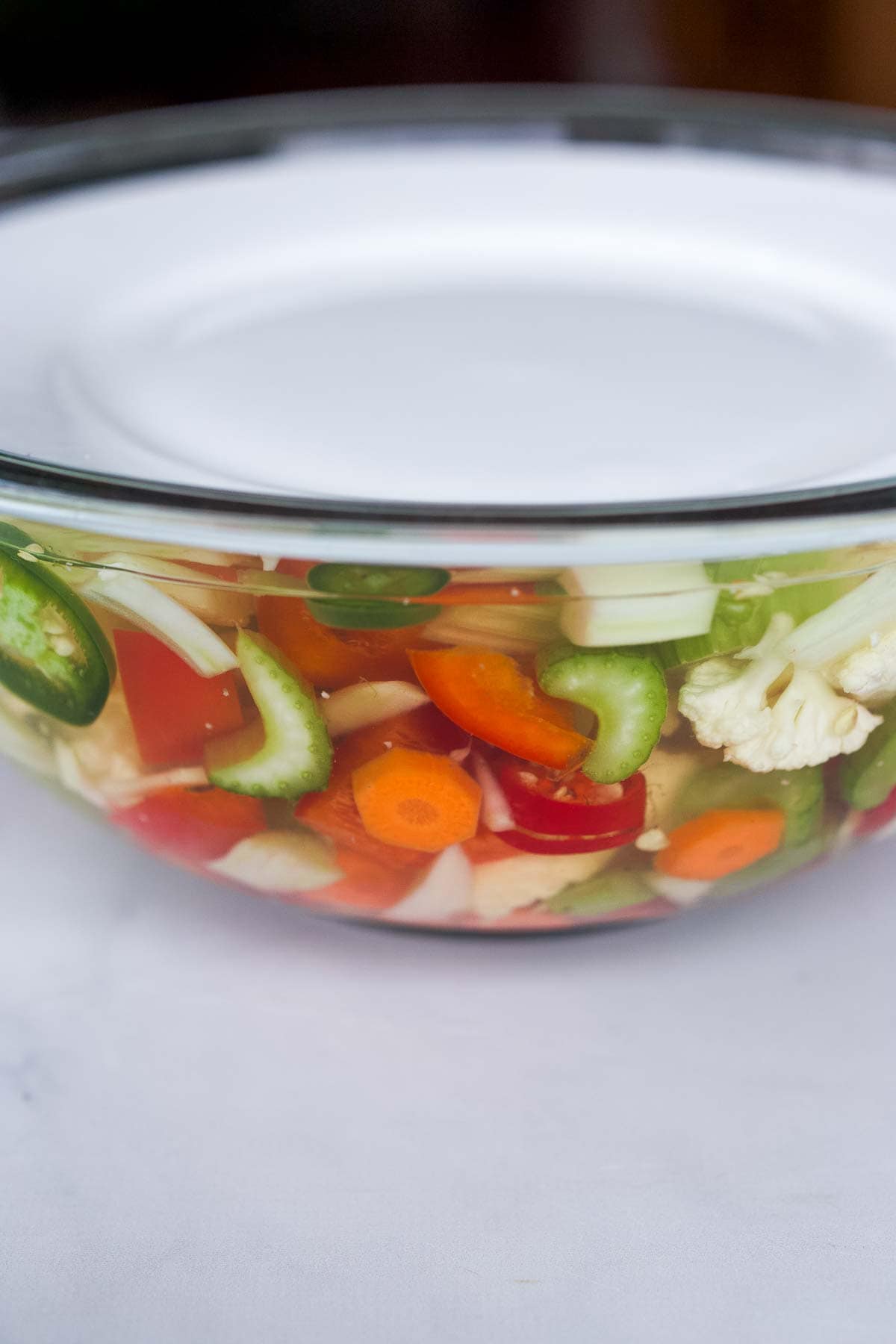 glass mixing bowl with giardiniera veggies covered in water with plate on top. 