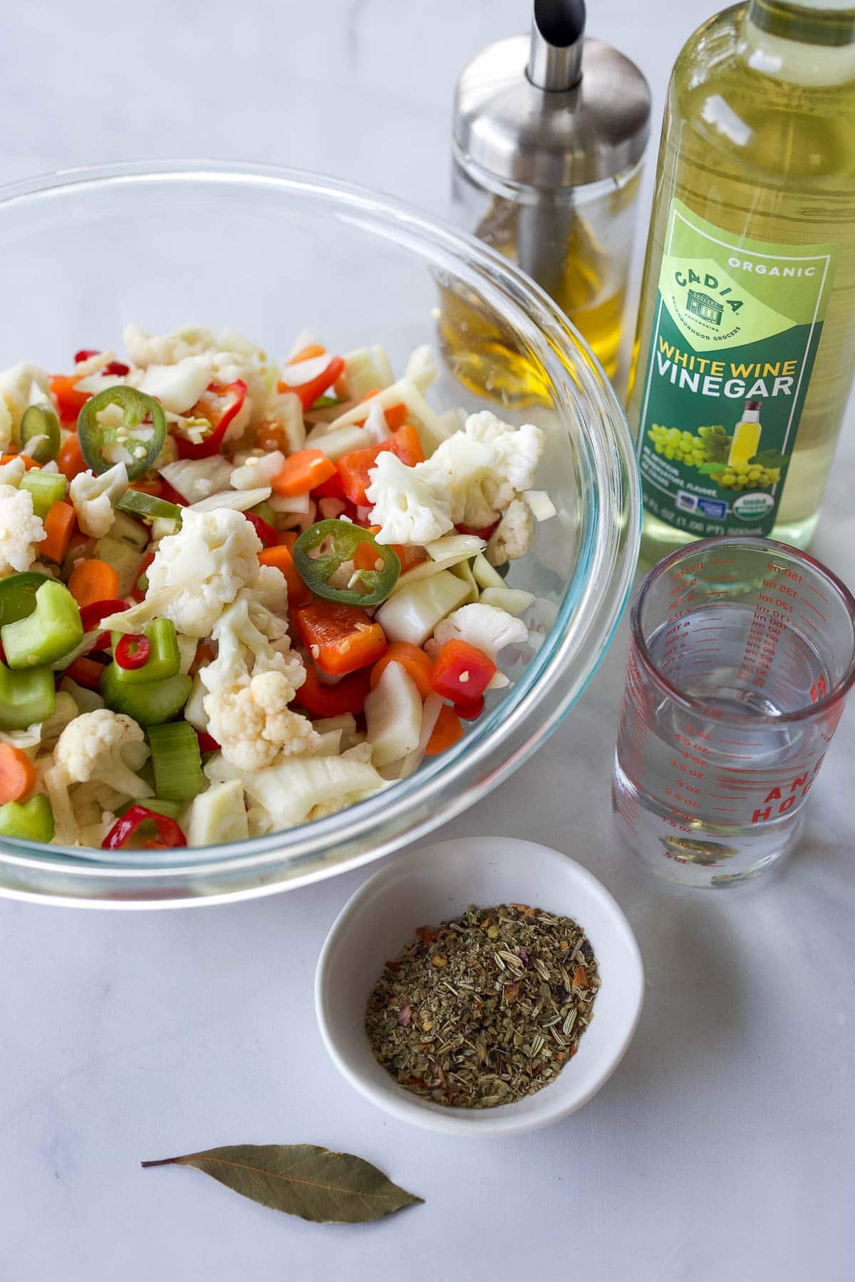 glass mixing bowl with giardiniera veggies next to oil, vinegar, water, spices, and bay leaf. 