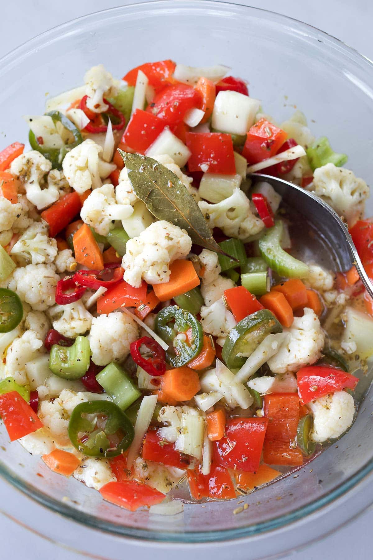 glass mixing bowl with spoon, with giardiniera - cauliflower, carrots, peppers, celery, fennel bulb coated in spices with a bay leaf. 