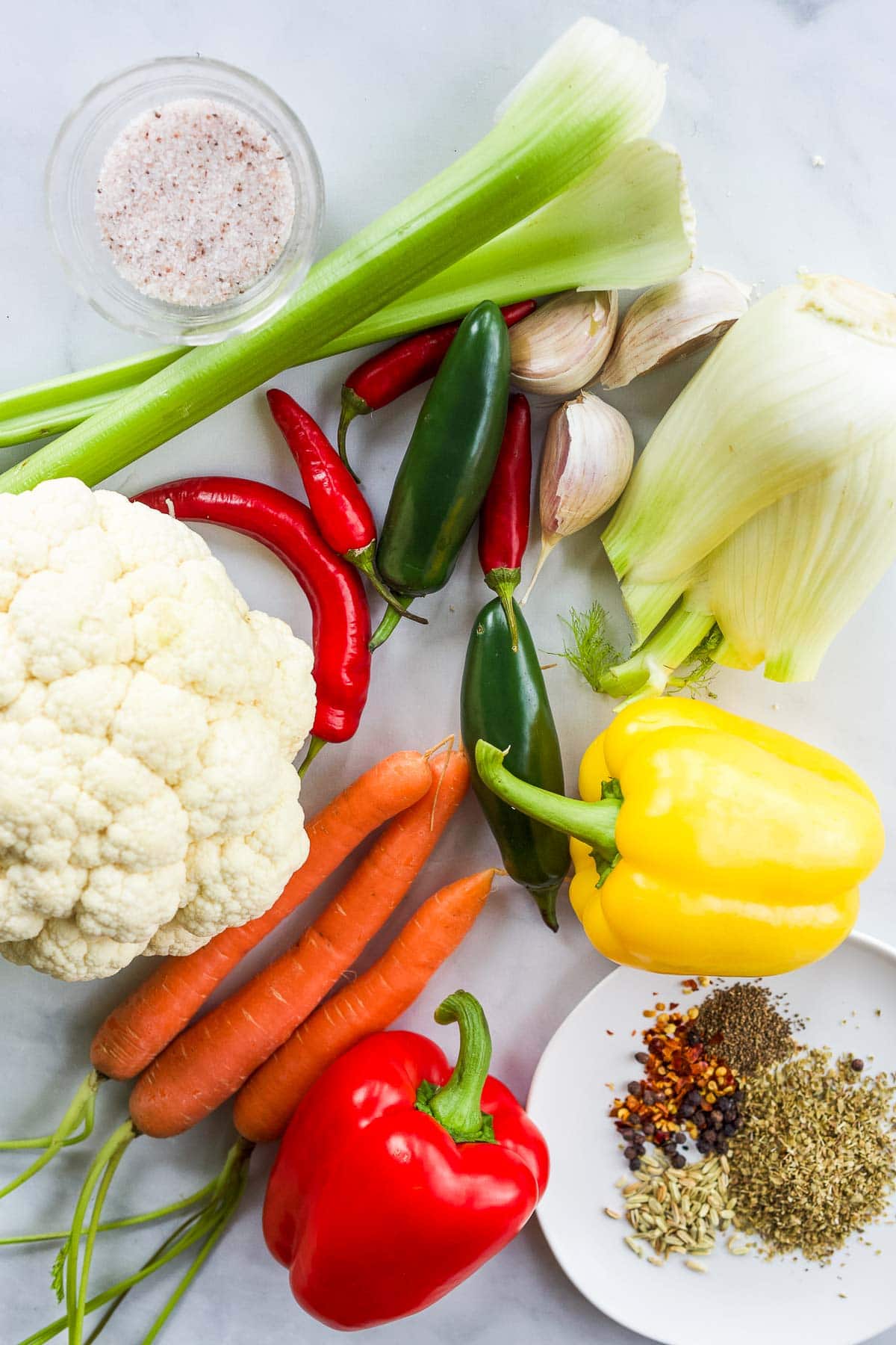ingredients laid out to make giardiniera - salt, celery, chilis, jalapeños, garlic, fennel bulb, bell peppers, cauliflower, carrots, bell peppers, spices. 