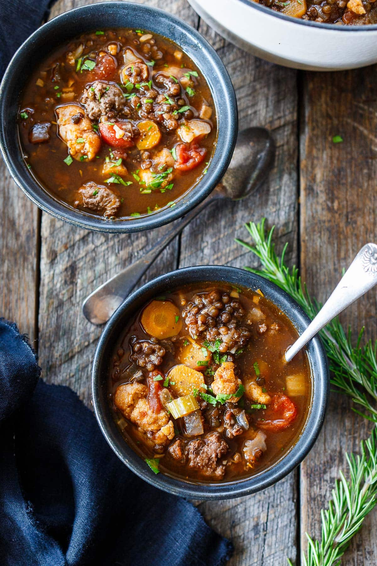 two bowls of lentil turkey sausage soup with spoons on wood surface next to sprigs of rosemary.