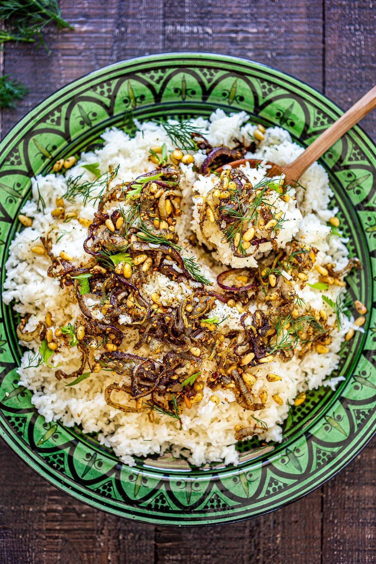 green serving dish with middle eastern rice topped with shallots, pine nuts, and fresh herbs, and a wood spoon holding up rice. 