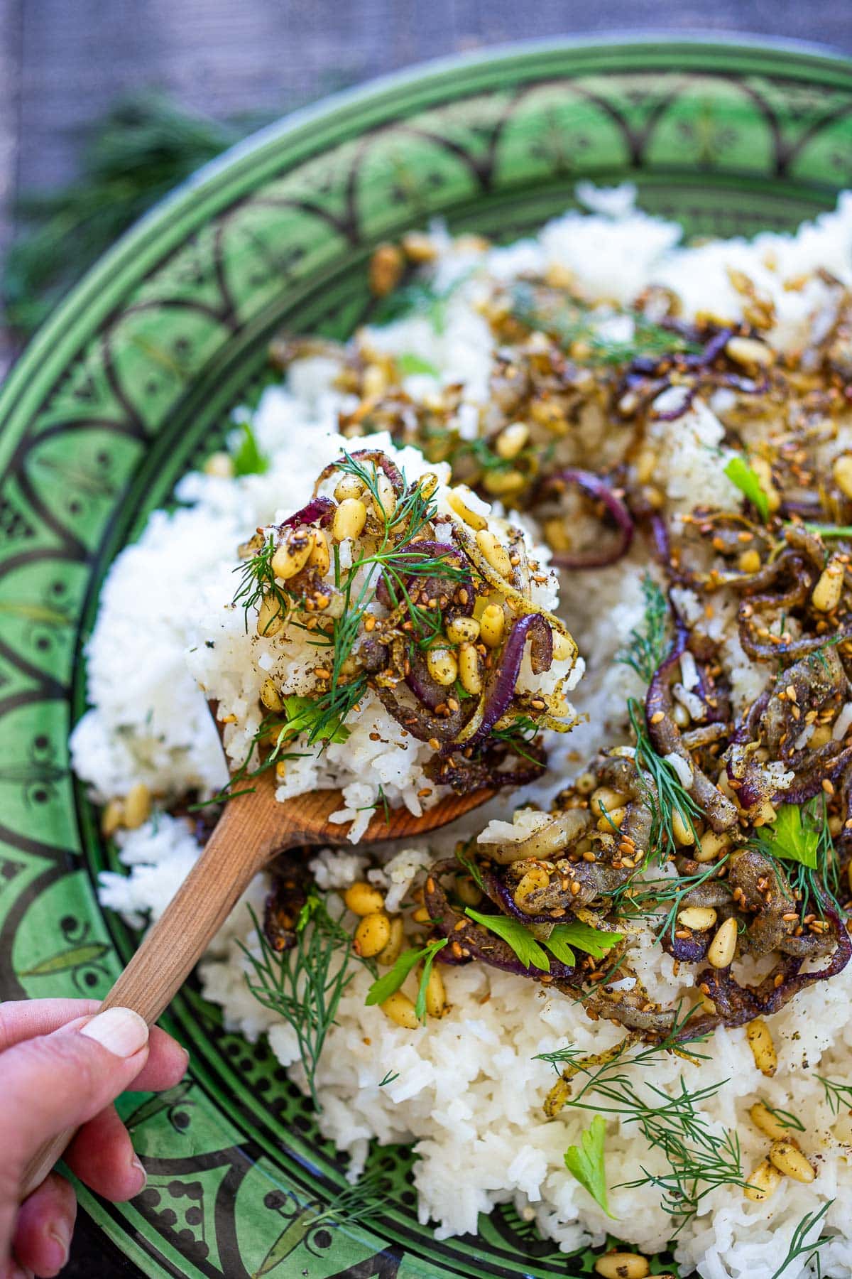 hand holding wood spoon full of middle eastern rice above serving dish topped with shallots, lemon zest, pine nuts, and herbs. 
