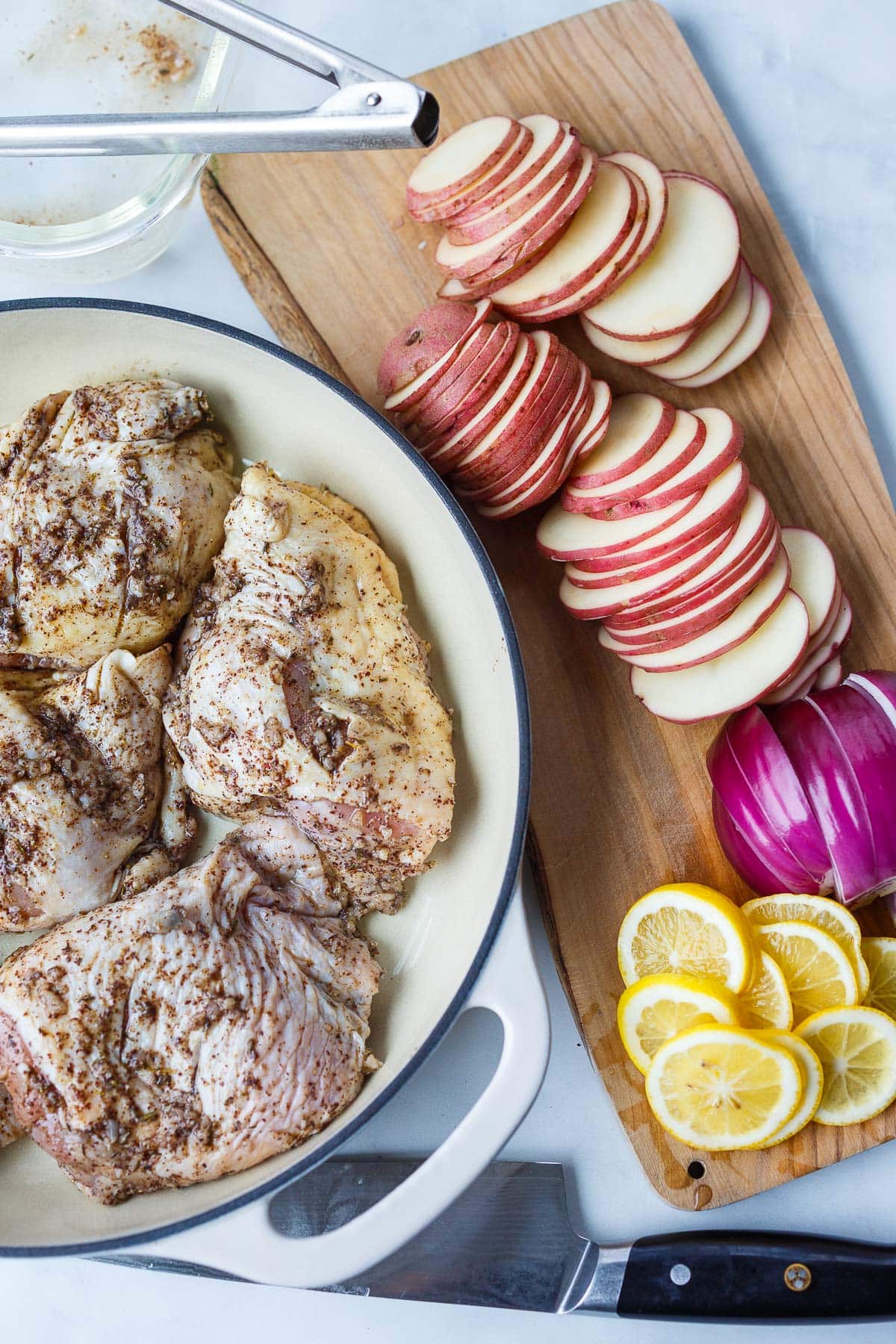 Wood board with sliced potatoes, red onions, and lemons next to dutch oven with marinated chicken thighs.