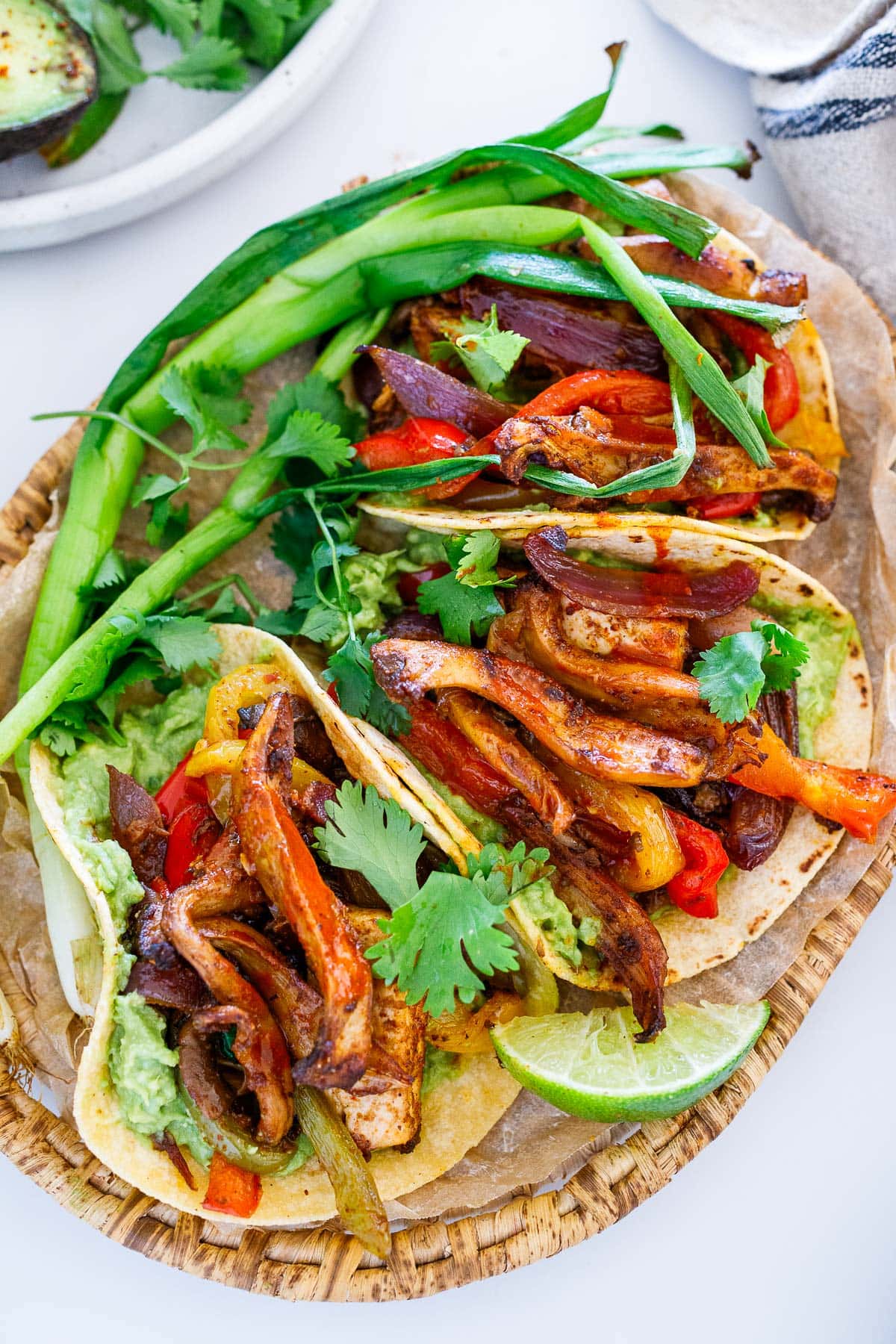 three tacos with vegan fajitas and mashed avocado topped with cilantro and green onion, served with lime wedge. 