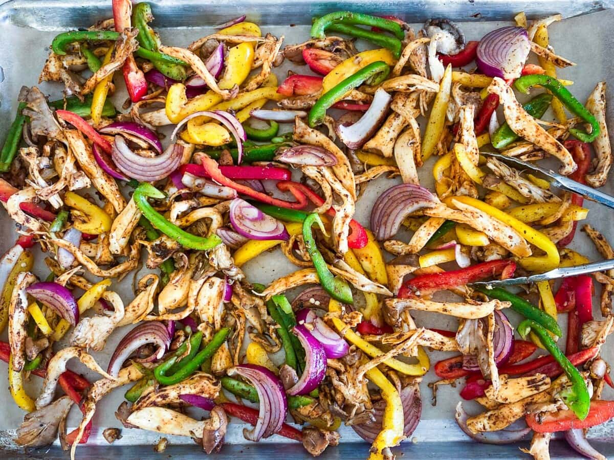 parchment lined sheet pan and tongs with sliced fajita veggies in marinade spread out.