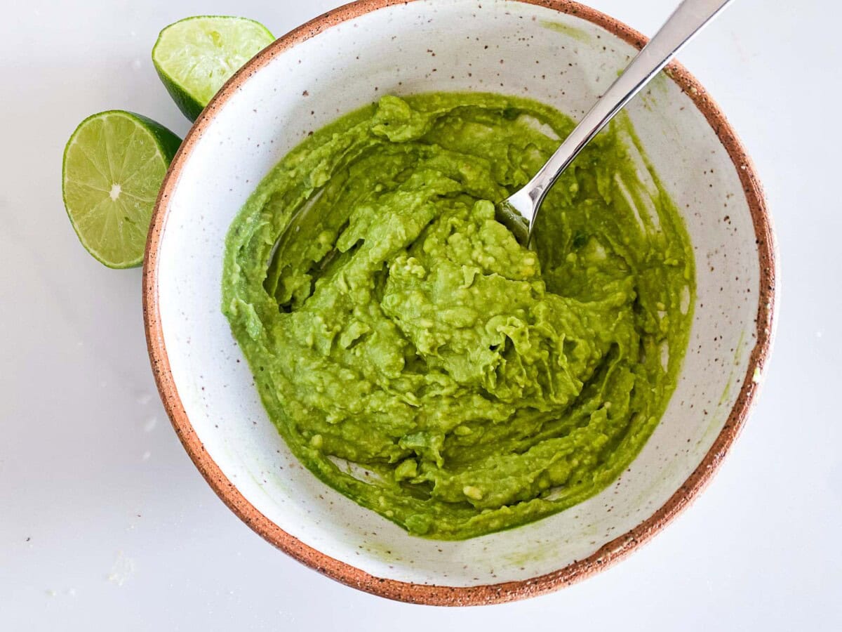 mashed avocado in bowl with fork next to lime halves. 