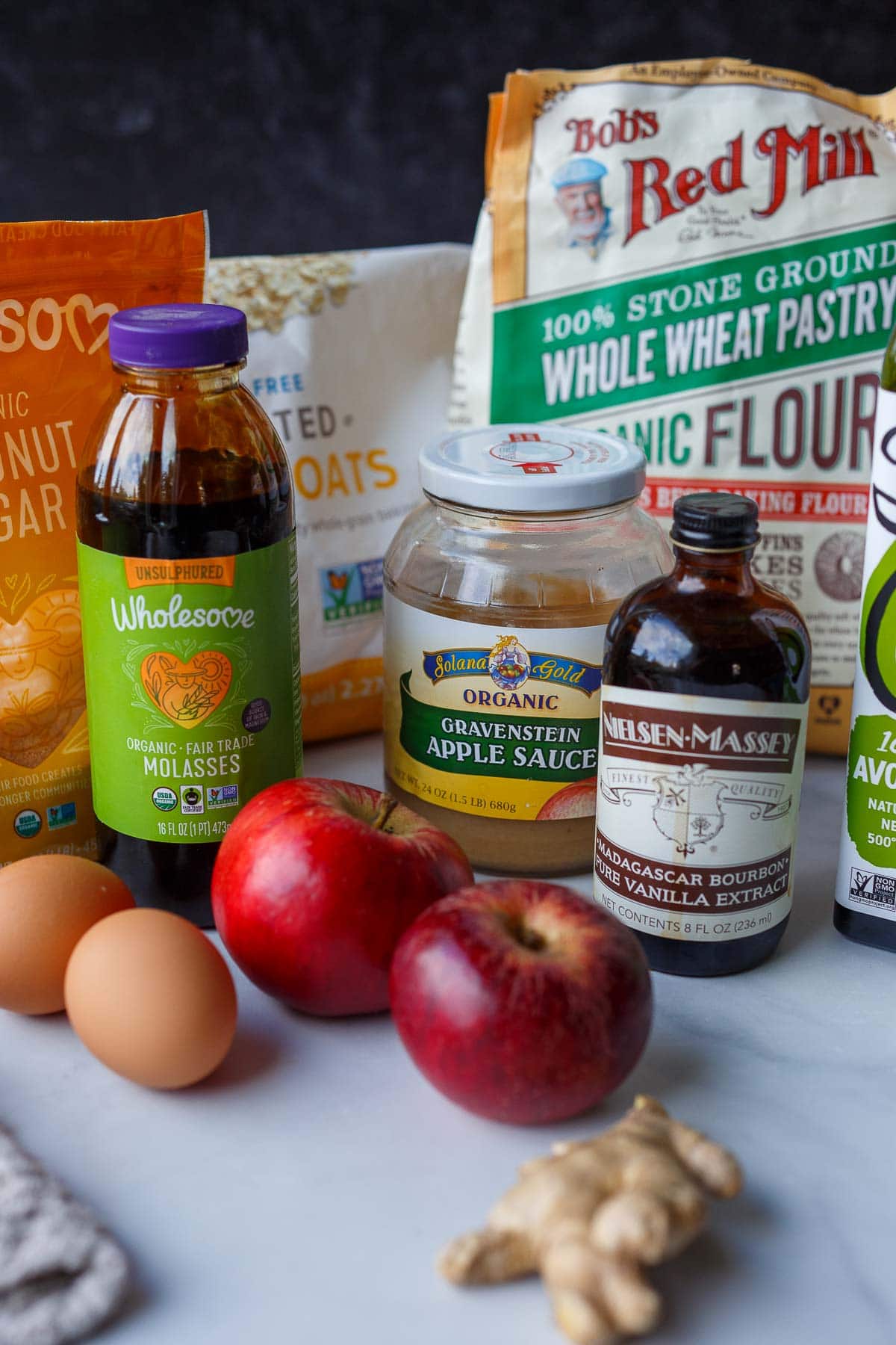 ingredients for apple oat muffins laid out on counter - whole wheat pastry flour, oats, coconut sugar, molasses, apple sauce, vanilla, avocado oil, apples, eggs, and ginger.