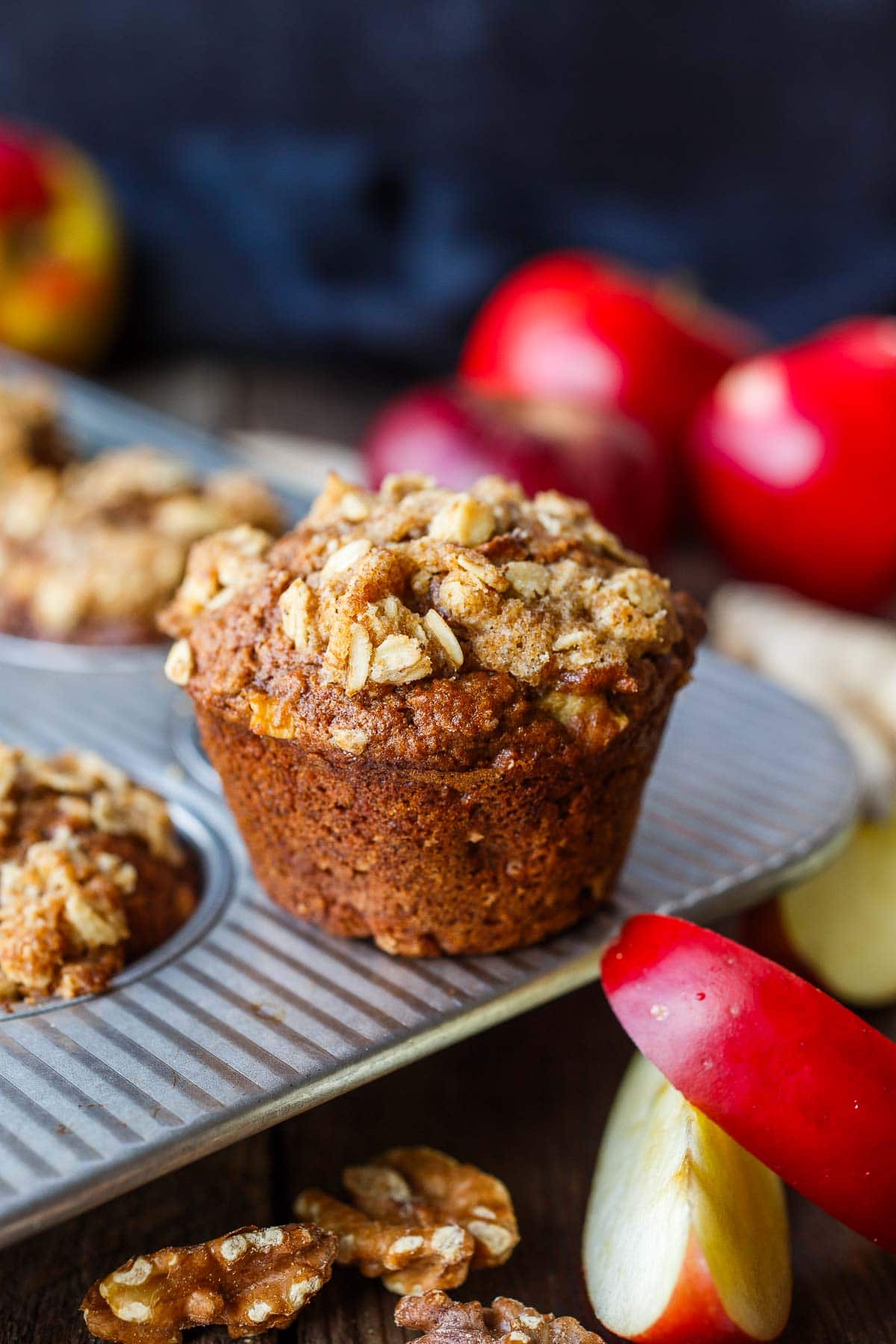 apple oat muffin with crumble topping standing on muffin tin surrounded by apples.