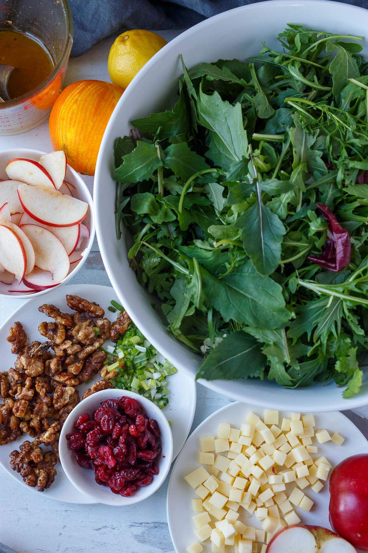 ingredients arranged for apple cheddar salad - bowl of mixed greens, orange and lemon, jar of dressing, small bowl with sliced apples, plate of cinnamon walnuts, dried cranberries, and sliced scallions, plate of cubed cheddar.
