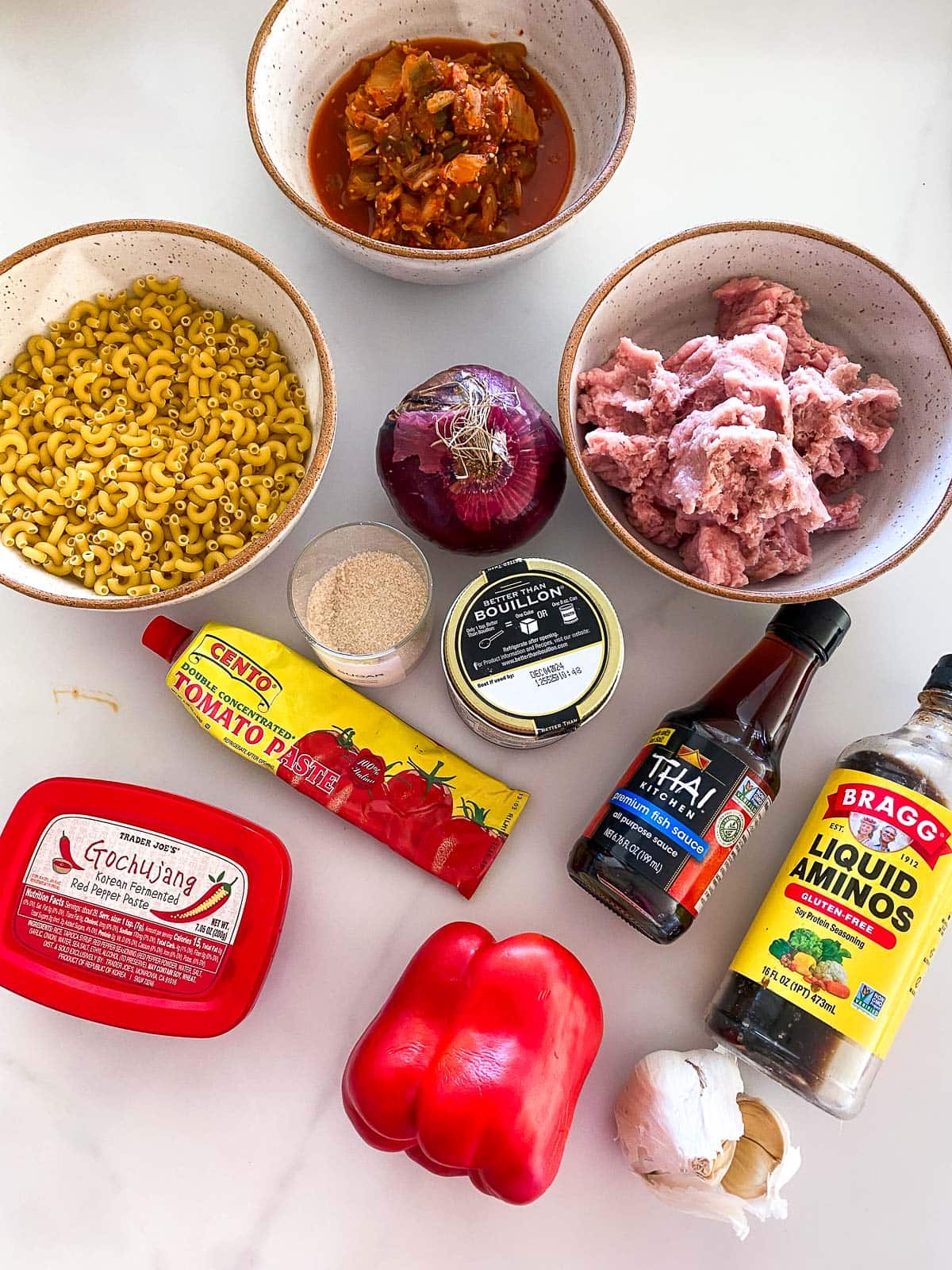 ingredients organized for kimchi chili mac and cheese - bowls of kimchi, macaroni, ground turkey, and red onion, sugar, bouillon, tomato paste, fish sauce, liquid aminos, gochujang, red bell pepper, garlic.