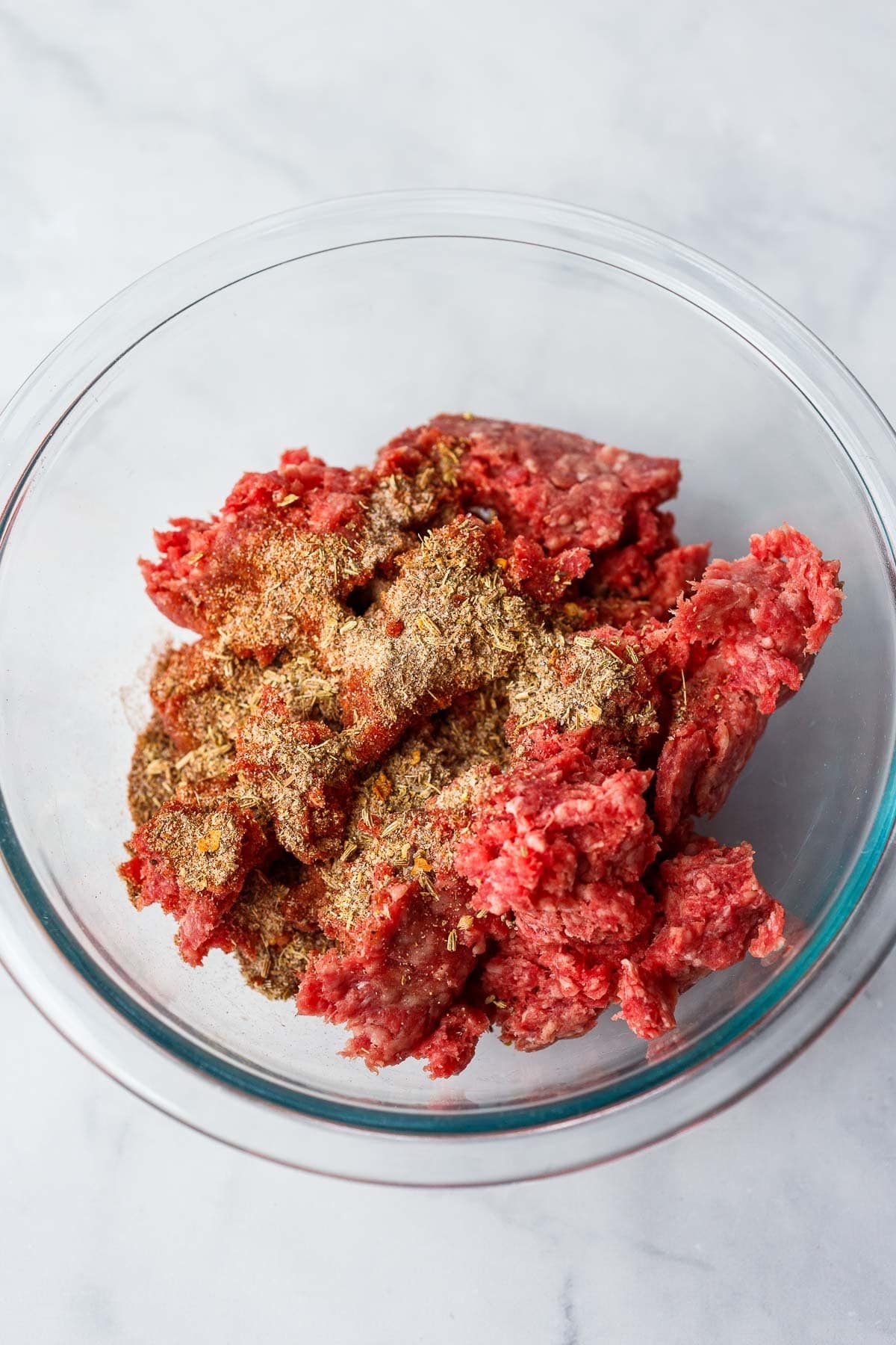 Italian seasoning and ground meat in glass mixing bowl. 